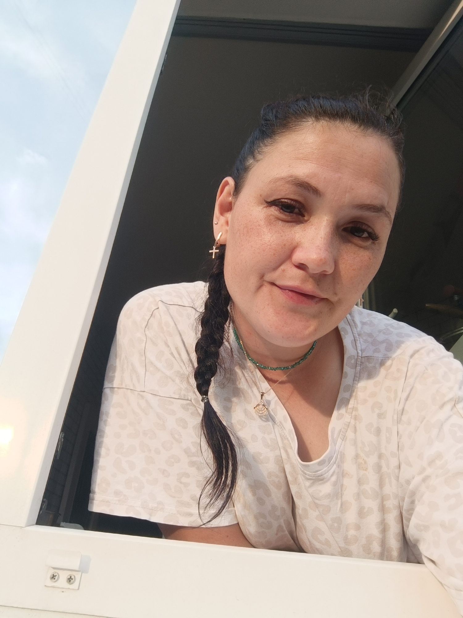 A medium shot captures a woman with dark hair pulled back in a braid, wearing a light-colored shirt with a leopard-print pattern. She is looking towards the camera with a slight smile and has freckles across her nose and cheeks. She is wearing a gold cross earring and a green beaded necklace with a gold pendant. The woman appears to be indoors, leaning on a white surface, possibly a windowsill or balcony railing, with a view of the sky and some architectural elements behind her. The lighting suggests it may be daytime. The background includes a dark area, possibly an interior space or a shadowed outdoor area, and a bright sky with some clouds.