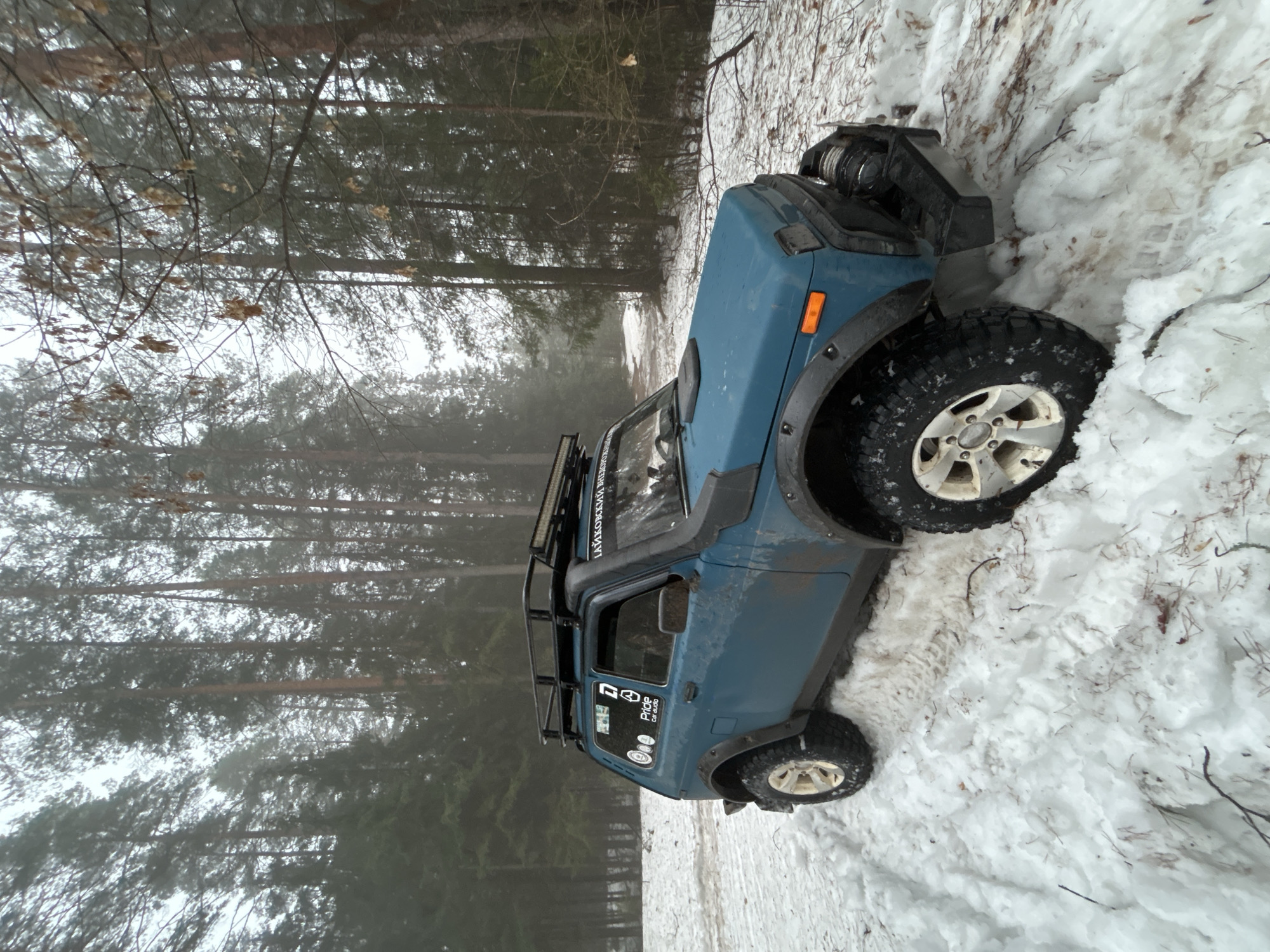 A blue Suzuki Samurai (or similar small SUV) is stuck in deep snow at the edge of a forest. The vehicle is angled slightly, with its front right wheel deeply embedded in the snowdrift. The surroundings are snowy with visible forest trees. Although the prompt mentions Chaikovsky, Russia, there are no visible details to confirm the location.