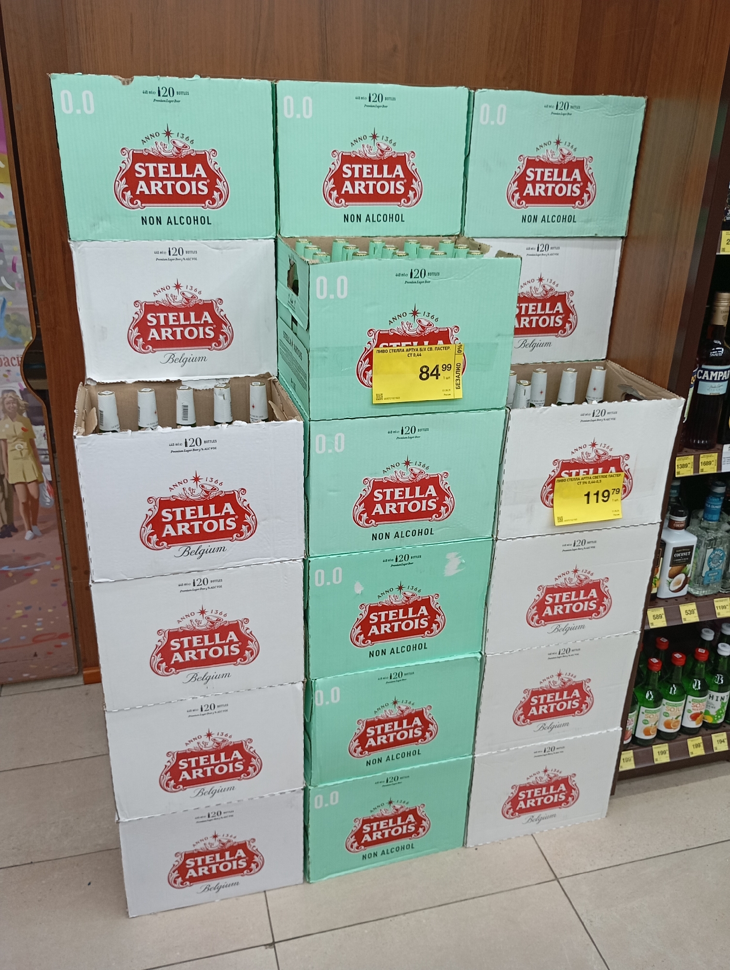 The image shows a retail display of Stella Artois beer bottles in a store. The display consists of multiple stacked boxes of the product. The boxes are predominantly white and light green with the Stella Artois branding. Some boxes also feature price tags with Cyrillic text. On the far left, a partial view of a poster or advertisement is visible, featuring a woman in a yellow outfit. To the right of the beer display, shelves stocked with other beverages, including bottles with green labels and various other brands, are visible. The flooring appears to be tiled. The lighting suggests an indoor retail environment. The presence of Cyrillic text on the price tags indicates the location is likely in a Russian-speaking country. Given the retail setting and the merchandise, it is plausible this scene is in Moscow, Russia.