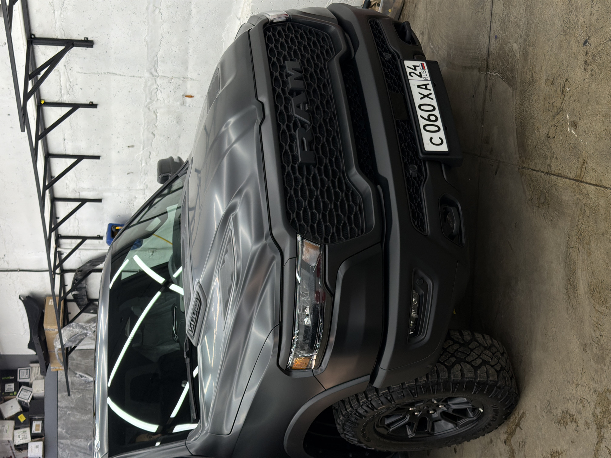 An eye-level shot captures the front and side of a large, matte dark grey or black Ram pickup truck, appearing to be a 1500 series with an "SST" badge visible on the side fender. The truck is positioned indoors, likely in a garage, workshop, or detailing bay, with the image rotated 90 degrees clockwise.
The truck features a prominent black honeycomb grille with "RAM" lettering emblazoned across it. Its headlights are angular and modern, and it is fitted with robust off-road tires and black multi-spoke wheels. A Russian license plate, "C 060 XA 24 RUS," is mounted on the front bumper. The "24 RUS" region code is consistent with Krasnoyarsk, Russia.
The surrounding environment consists of textured, light-colored concrete or painted walls, showing some signs of wear and staining. To the left of the truck, a black metal shelving or support structure is visible, along with stacks of cardboard boxes and other items, suggesting a storage or work area. The concrete floor beneath the truck is also textured and shows marks. Linear artificial lights are reflected in the truck's windshield and hood, illuminating the vehicle and indicating an indoor setting. No people are visible, and the truck appears stationary, possibly undergoing maintenance or preparation.