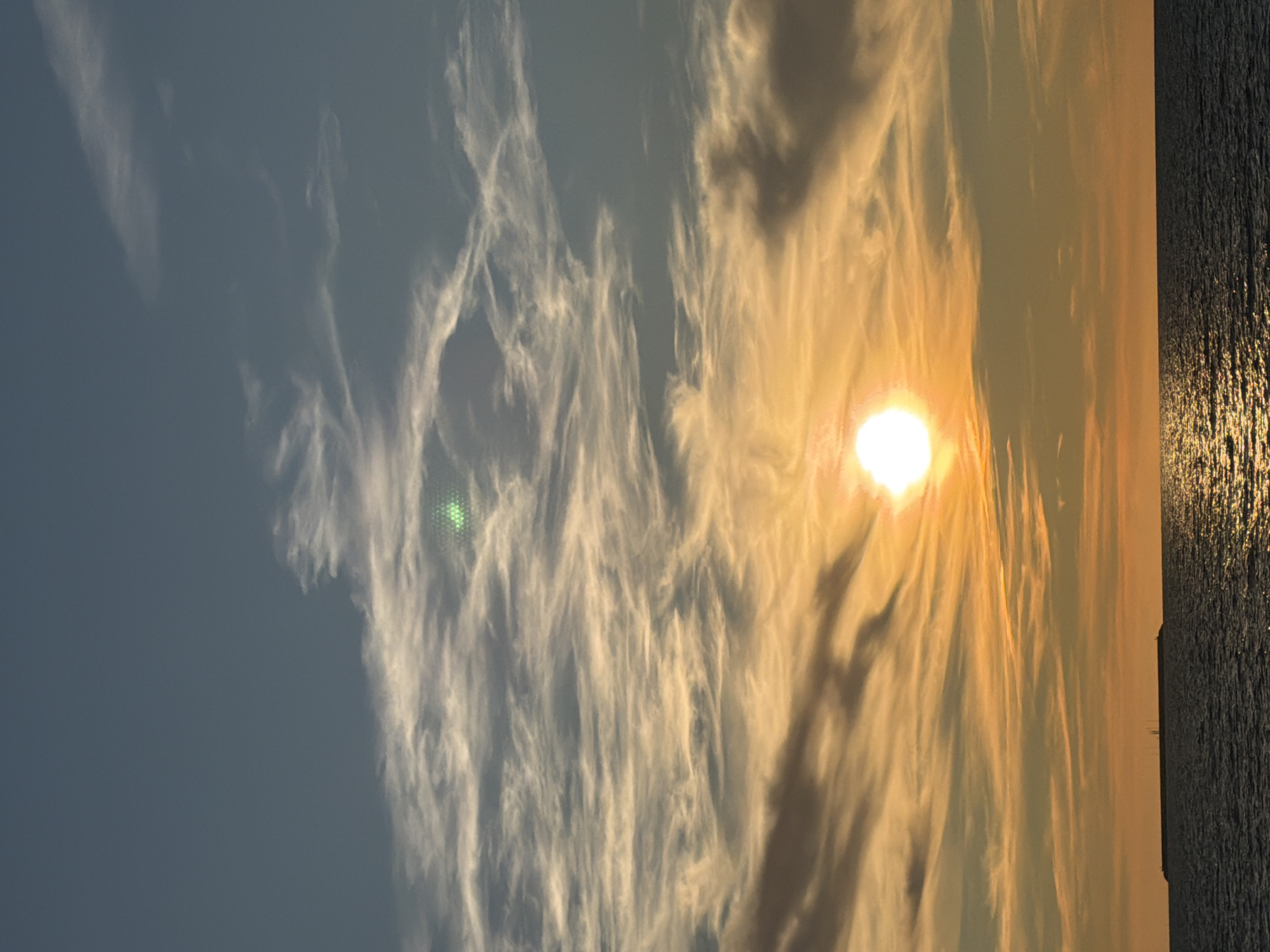 The image depicts a sunset over the Baltic Sea, with a sliver of land visible on the horizon. The sky is a gradient of colors, from a pale blue at the top to vibrant oranges and yellows closer to the horizon, illuminated by the setting sun. Wispy clouds streak across the sky, catching the warm light. The sea is dark and choppy, with reflections of the sunset shimmering on its surface. On the distant landmass, several small structures, possibly lights or antennas, are silhouetted against the sky. The scene appears serene and natural, with no people or specific man-made objects immediately apparent other than the distant structures and what might be a breakwater. The presence of a lighthouse or similar navigational aid is suggested by the solitary tower-like structure on the horizon. Based on the characteristics of the sky, clouds, and the sea, this could be a typical sunset scene observed from the coast of Zelenogradsk, Russia.