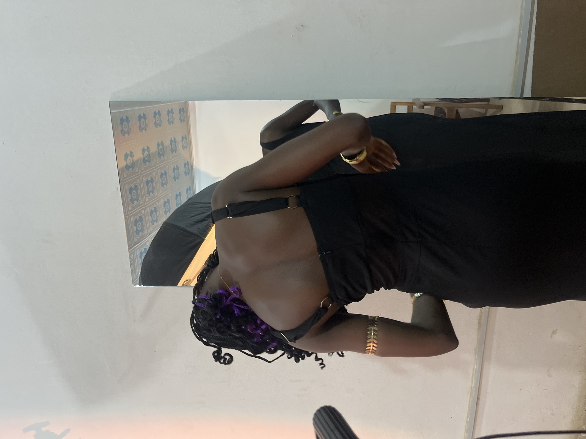 In Gura Top, Nigeria, a person with dark skin and braided hair adorned with purple extensions is standing with their back to the camera, facing a large mirror. They are wearing a black sleeveless dress and a gold arm cuff on their left bicep. The mirror reflects the person and a portion of the room, including a large black object, possibly a backdrop or lighting equipment, and a tiled ceiling with a floral pattern. The person appears to be adjusting their dress or hair, leaning forward slightly. The environment is a room with white walls. A portion of a microphone stand is visible on the left side of the image.