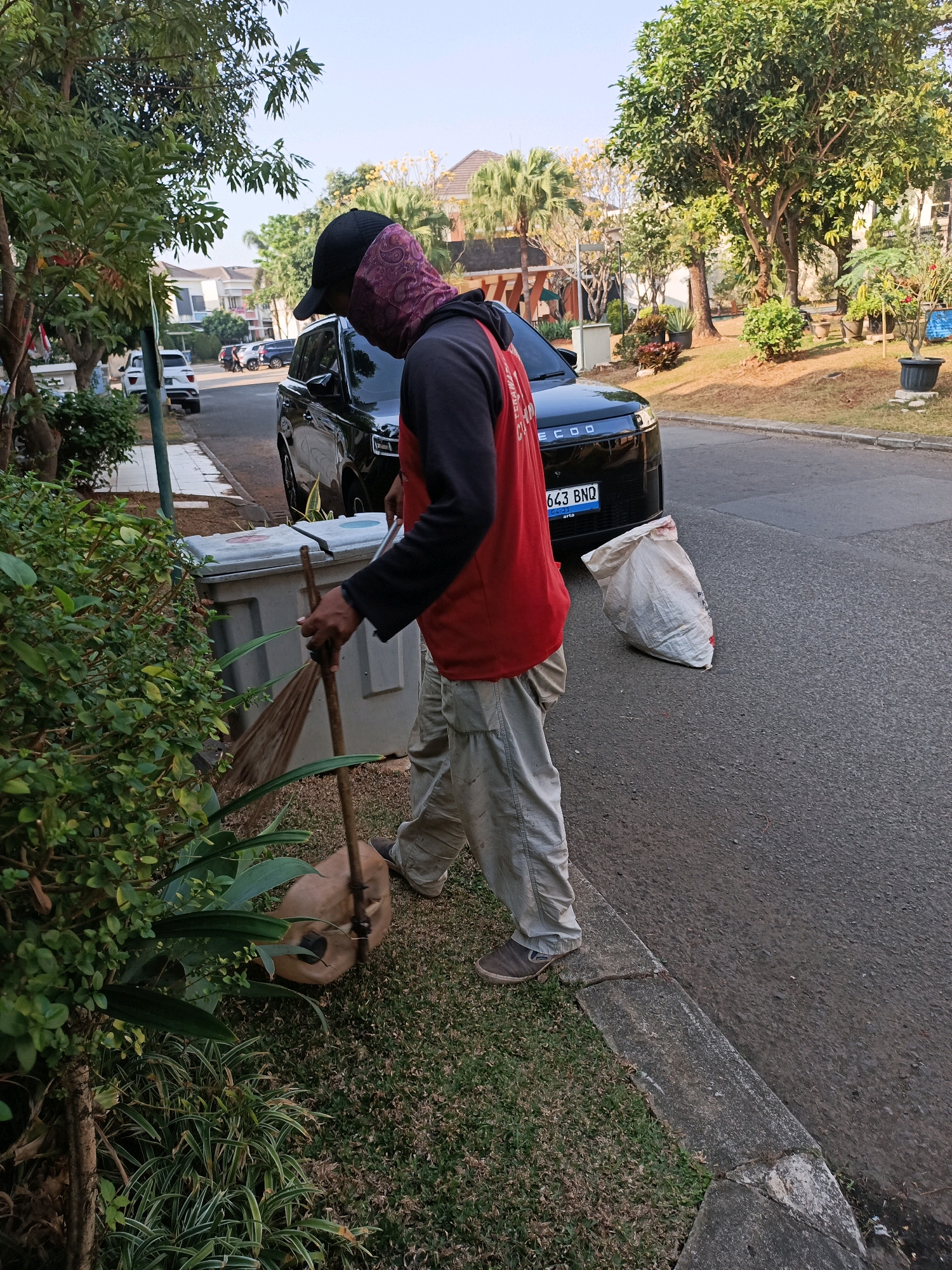 A person is sweeping a grassy area next to a road in Curug Sangereng, Indonesia. The individual, wearing a black baseball cap, a red long-sleeved shirt, and tan pants, is using a broom to gather debris. A black SUV is parked on the road to the right of the person, with a white trash bag visible behind it. In the background, there are houses and trees, suggesting a residential neighborhood. The scene is lit by natural sunlight, indicating it is daytime.