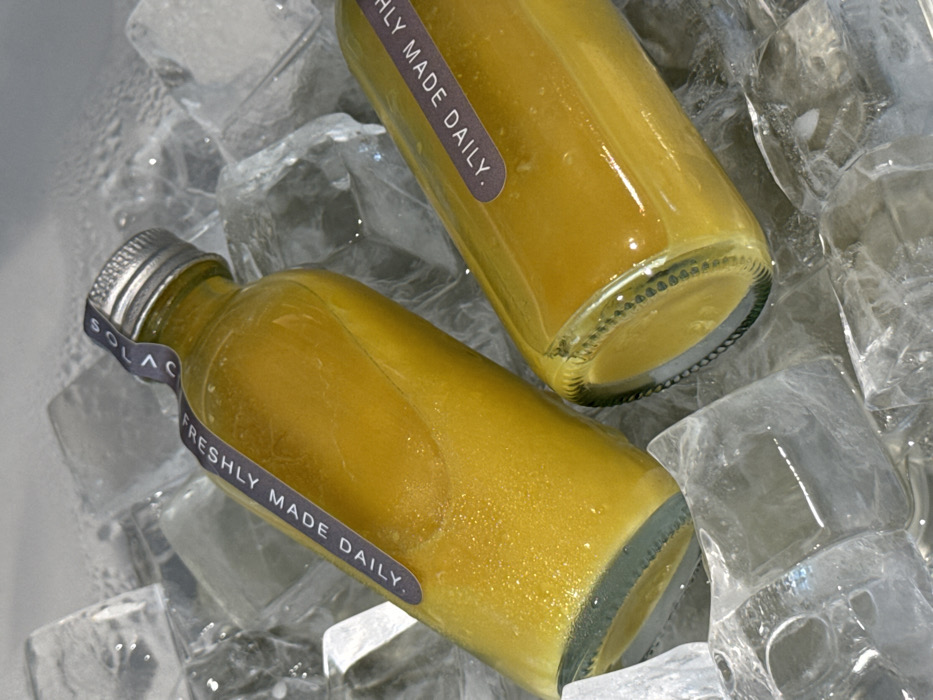 Two glass bottles filled with yellow liquid, possibly juice, rest on a bed of ice. A label reading "FRESHLY MADE DAILY" wraps around each bottle. One bottle displays a silver cap with "SOLACE" visible on a sticker. The clear ice cubes reflect light, creating highlights on the bottles and surrounding surfaces. The close-up shot emphasizes the chilled and refreshing nature of the beverage.