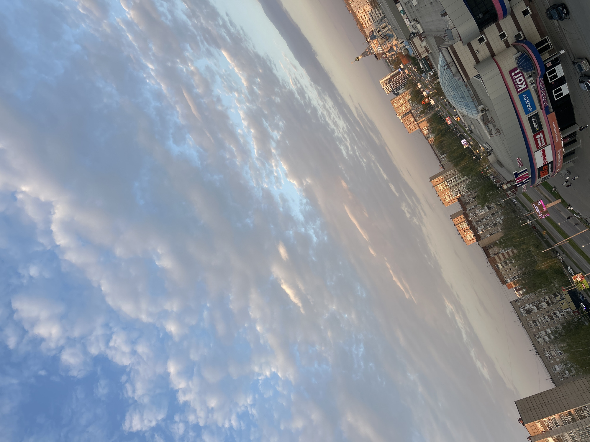 The image shows a view of Izhevsk, Russia, featuring a shopping center with visible signs for "kari" and "SOKOLOV." Buildings and streets extend into the distance. Above, there's a view of a cloudy sky, with blue peeking through.