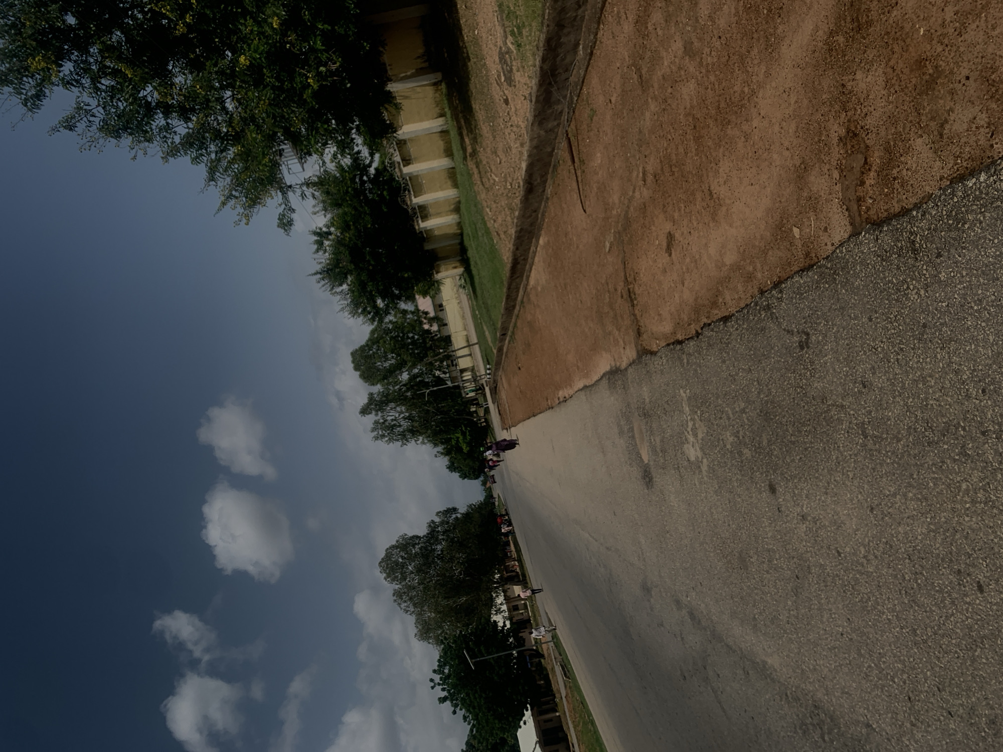 The image depicts a street scene in Jalingo, Nigeria, under a partly cloudy sky. The perspective is from street level, angled slightly upward. On the left side of the road, buildings with light-colored walls and dark roofs are visible, interspersed with trees and a solar-powered street light. Several people are walking on the sidewalk and on the road. The road itself is paved and shows some wear. On the right side of the road, a strip of reddish-brown dirt or sand runs alongside a low concrete barrier, separating it from a grassy area and a tall, yellow wall topped with barbed wire, suggesting a property boundary. Trees with dense green foliage and some yellow flowers line the area behind the wall. Several cars are visible in the distance on the road. The overall impression is of a developing urban or semi-urban environment.