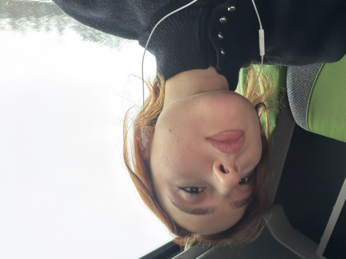 The image depicts a young light-skinned female in a low-angle close-up within a vehicle, located in Moineville, France. She has reddish-brown hair, styled loosely, and light makeup on her eyes and lips. She wears a black high-neck top with several silver-toned snap buttons visible on the collar area. White wired earphones are inserted into both ears, with the wire draped across her left shoulder and chest. Her gaze is directed slightly upwards and to the viewer's right. To the left, behind her, a portion of a vehicle seat with green and grey upholstery is visible. To the right, through a window, an overcast sky is visible, with streaks of water indicating rain on the glass surface. The interior of the vehicle has dark overhead elements. No other individuals are present.