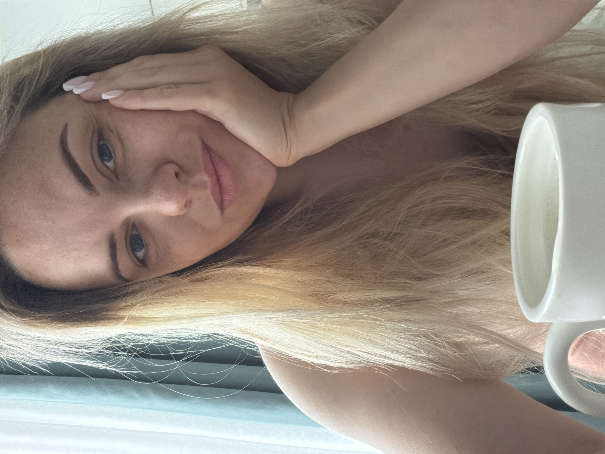 A woman with long, blonde hair is lying down, with her head slightly tilted. She is holding her hand to her face. There is a white mug in the right side of the image. The scene is indoors, but the location is not in Votkinsk, Russia as there is no indication in the photograph to suggest this.