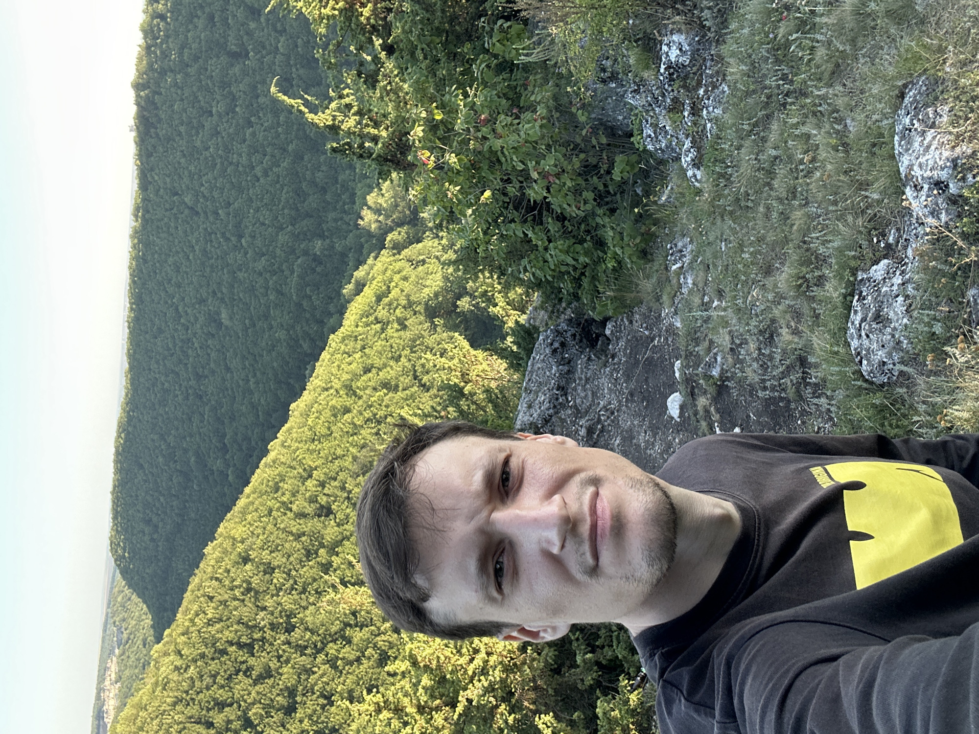A selfie captured from a high vantage point showcases a young man in the foreground, looking directly at the camera. He is wearing a dark long-sleeved shirt with a prominent yellow graphic on the left side of his chest. His facial features are clearly visible, with short brown hair and a slight stubble. The background reveals a sweeping landscape of densely forested hills, typical of the Makiv region in Ukraine, bathed in the soft light of what appears to be late afternoon or early morning. The terrain suggests a natural, somewhat rugged environment with rocky outcrops and grassy patches visible around the man. The distant horizon shows a clear sky with a faint hint of a settlement or town.