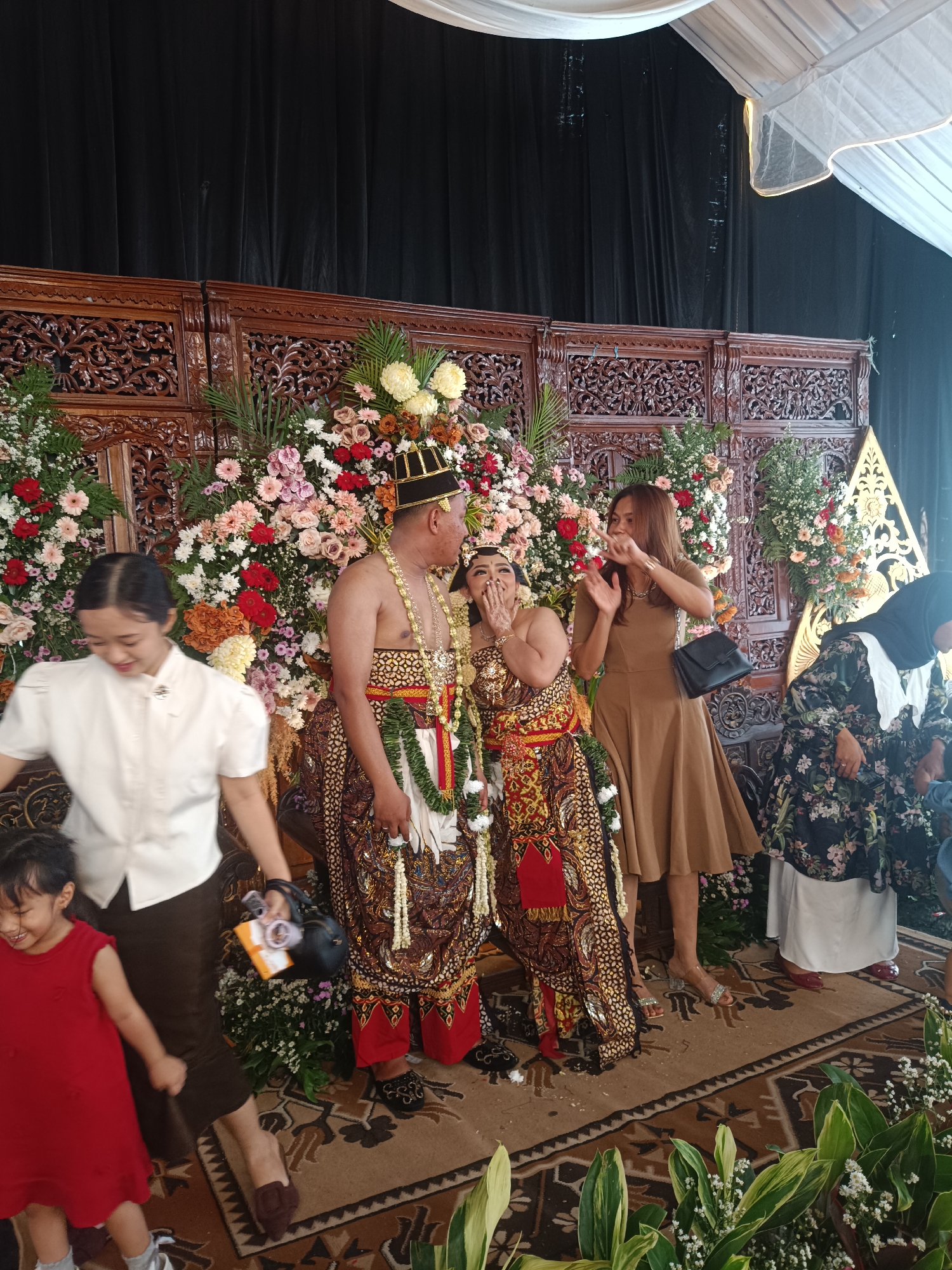 In Depok, Indonesia, at a wedding ceremony, a couple in traditional attire stands before a floral backdrop. The groom, shirtless and wearing a headdress and ornate waistcloth, stands to the left of the bride, who is adorned in similar traditional clothing. A woman in a tan dress gestures with her hands, while another woman in a floral dress and hijab is partially visible on the right. A young girl in a red dress stands to the left, and a woman in a white blouse is to her left, appearing to hand something to the bride. The environment is decorated with abundant flowers and intricate wooden carvings, suggesting a formal celebration.
