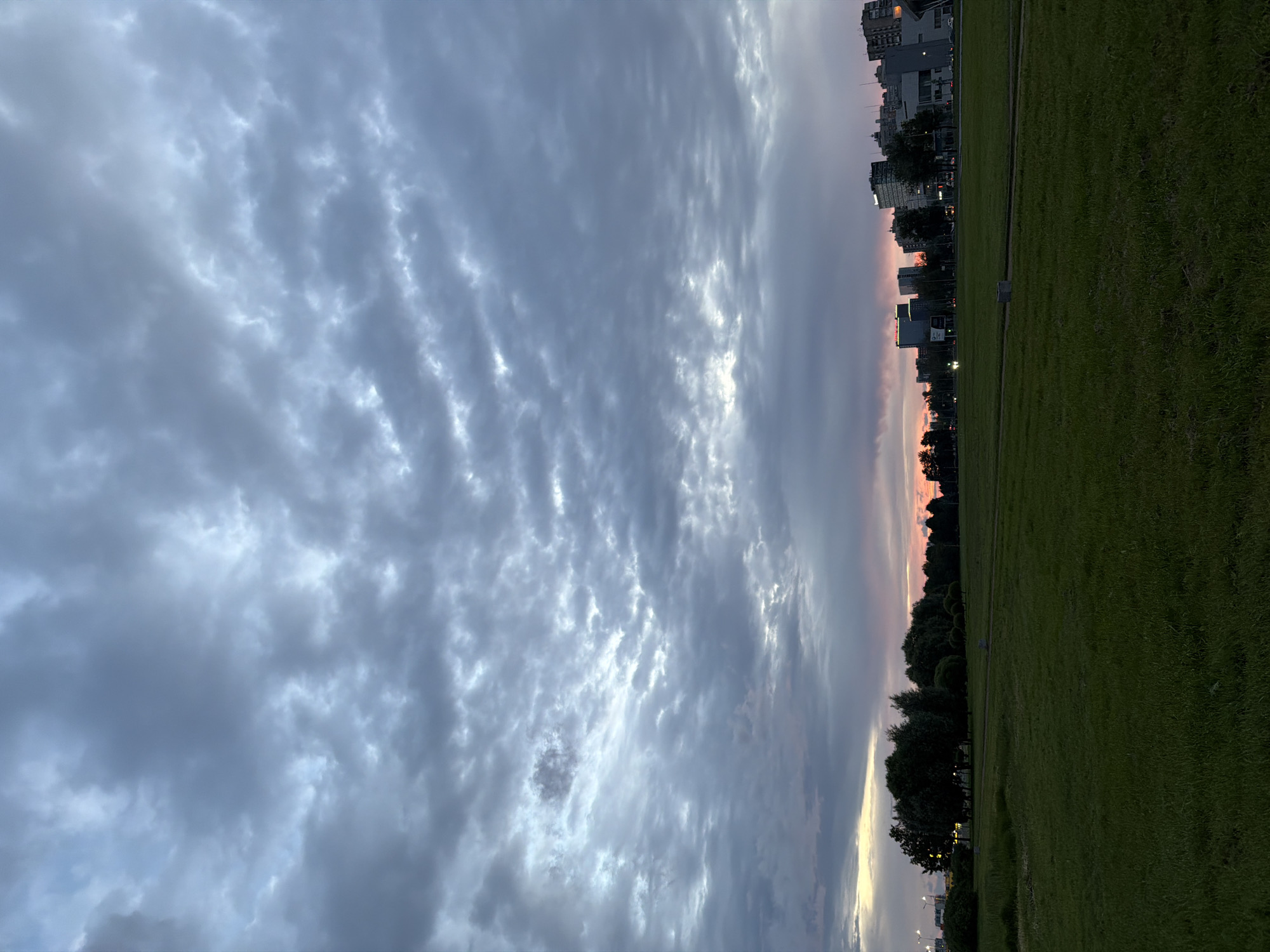 The image depicts a city skyline at dusk, with a large, grassy field in the foreground. The sky is filled with dramatic, grey clouds, tinged with pink and orange from the setting sun. A line of trees separates the field from the urban landscape, where various buildings of different heights and architectural styles are visible. Streetlights illuminate the roads and some buildings are lit from within. The field itself is dark green and appears to be well-maintained, with a path or road cutting through it. The overall impression is one of a peaceful, urban park at the end of the day.