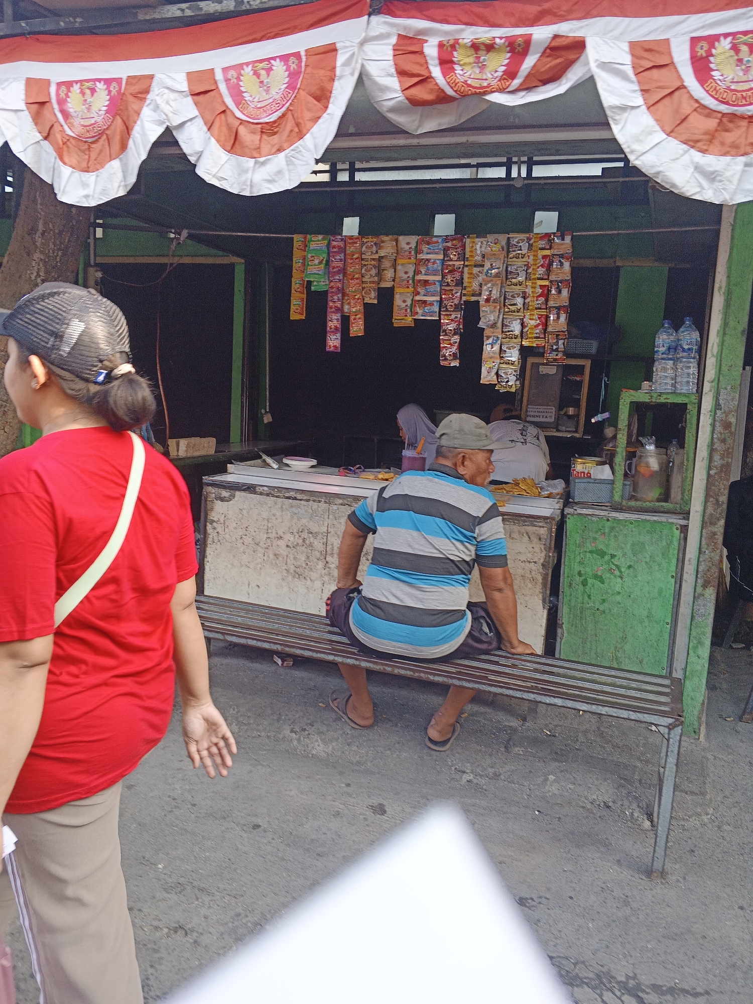 An outdoor scene in Krian, Indonesia depicts a small food stall adorned with Indonesian flags. A woman in a red shirt and a baseball cap is partially visible on the left, facing away from the camera. In the center, a man wearing a striped t-shirt and a baseball cap sits on a metal bench, facing the stall. The stall displays various packaged goods, including snacks and bottled water. A green metal cabinet is positioned to the right of the stall. The ground is paved with concrete.