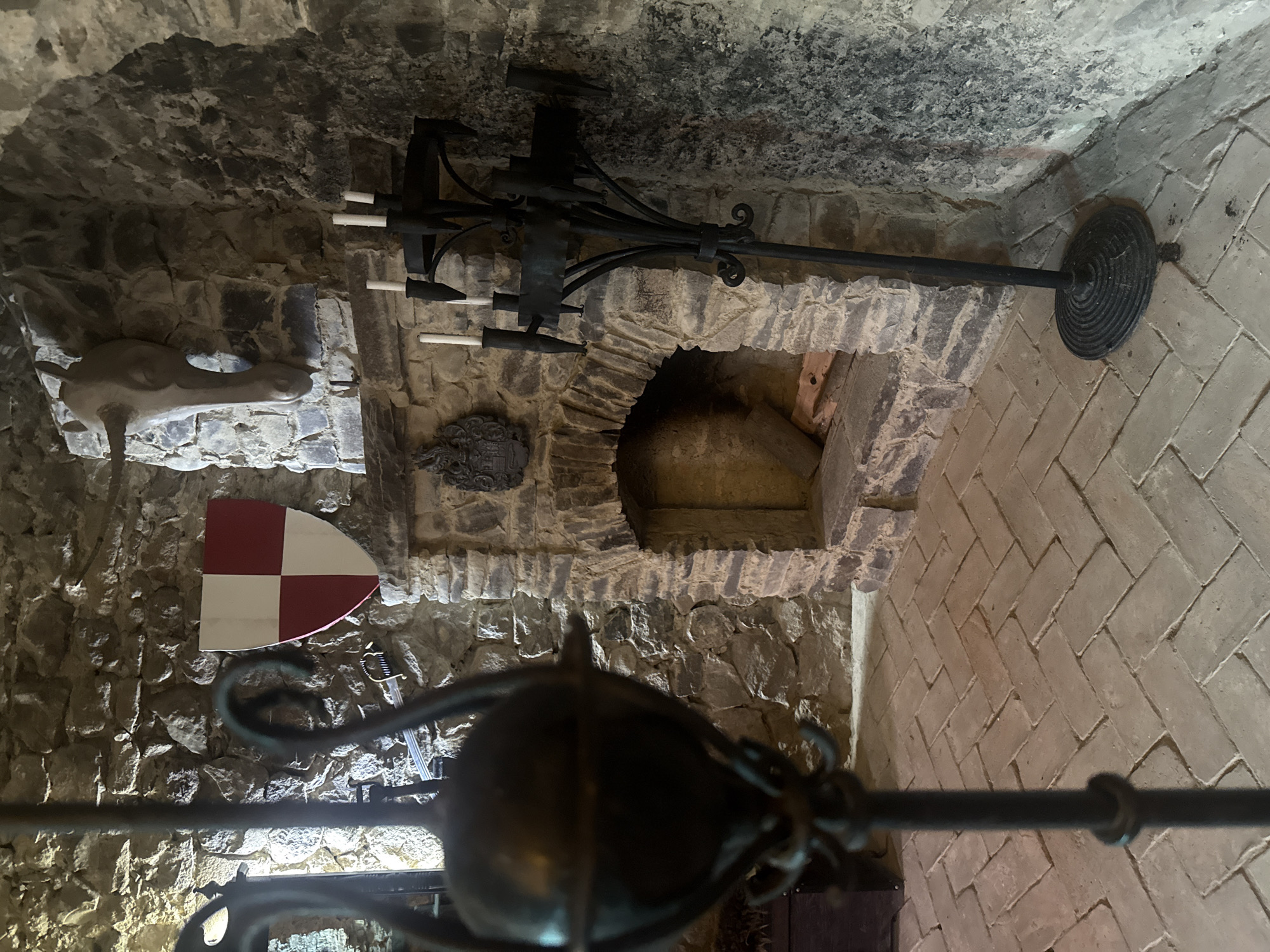 The image captures the interior of the Saint Miklós Castle in Chynadiiovo, Ukraine. The scene is an alcove within a stone wall, decorated with a coat of arms shield in red and white. Above it, there is a carved stone horse. The alcove features an arched opening, possibly leading to another part of the castle, and a large metal candelabra on a stand. The room is illuminated by an external light source. A secondary decorative metal candelabra is partially in the foreground. The walls are constructed of irregular stones, while the floor consists of rectangular bricks laid in a herringbone pattern.