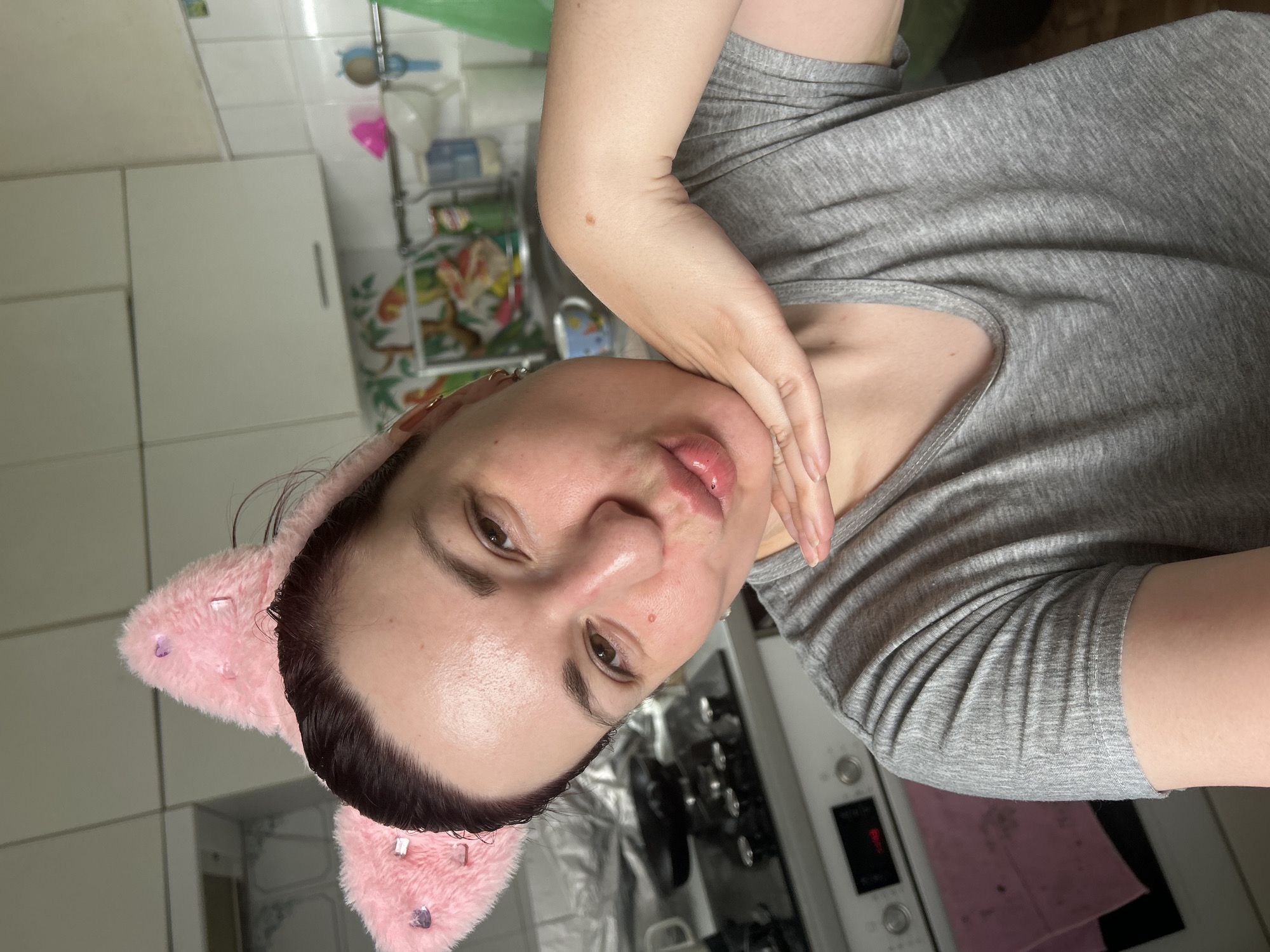 A young woman with dark hair and cat ears, possibly for a costume, leans forward, puckering her lips for a selfie. She is wearing a plain grey t-shirt and has her left hand resting on her chin. The background appears to be a kitchen, with white cabinets and a stovetop visible. There are no explicit indications of the city of Фоки, Russia, in the image.