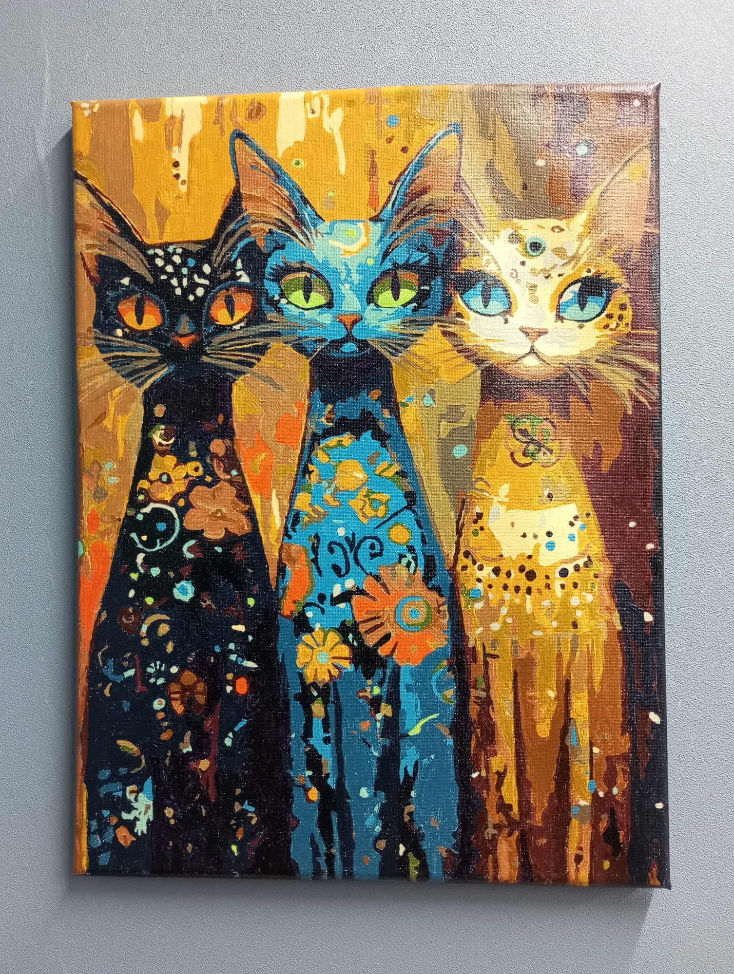 The image is a close-up of an oil painting of three stylized cats. The cats are arranged side-by-side, facing forward. The cat on the left is primarily black with orange eyes and decorative patterns on its body. The center cat is blue with green eyes and floral designs. The cat on the right is yellow with blue eyes and spotted patterns. The background is a textured blend of brown and yellow tones. The canvas is stretched on a frame, indicating it's a finished piece of art. The image is taken indoors against a plain gray wall.
There is no discernible connection to the city of Pervomaysky, Russia, in the image.