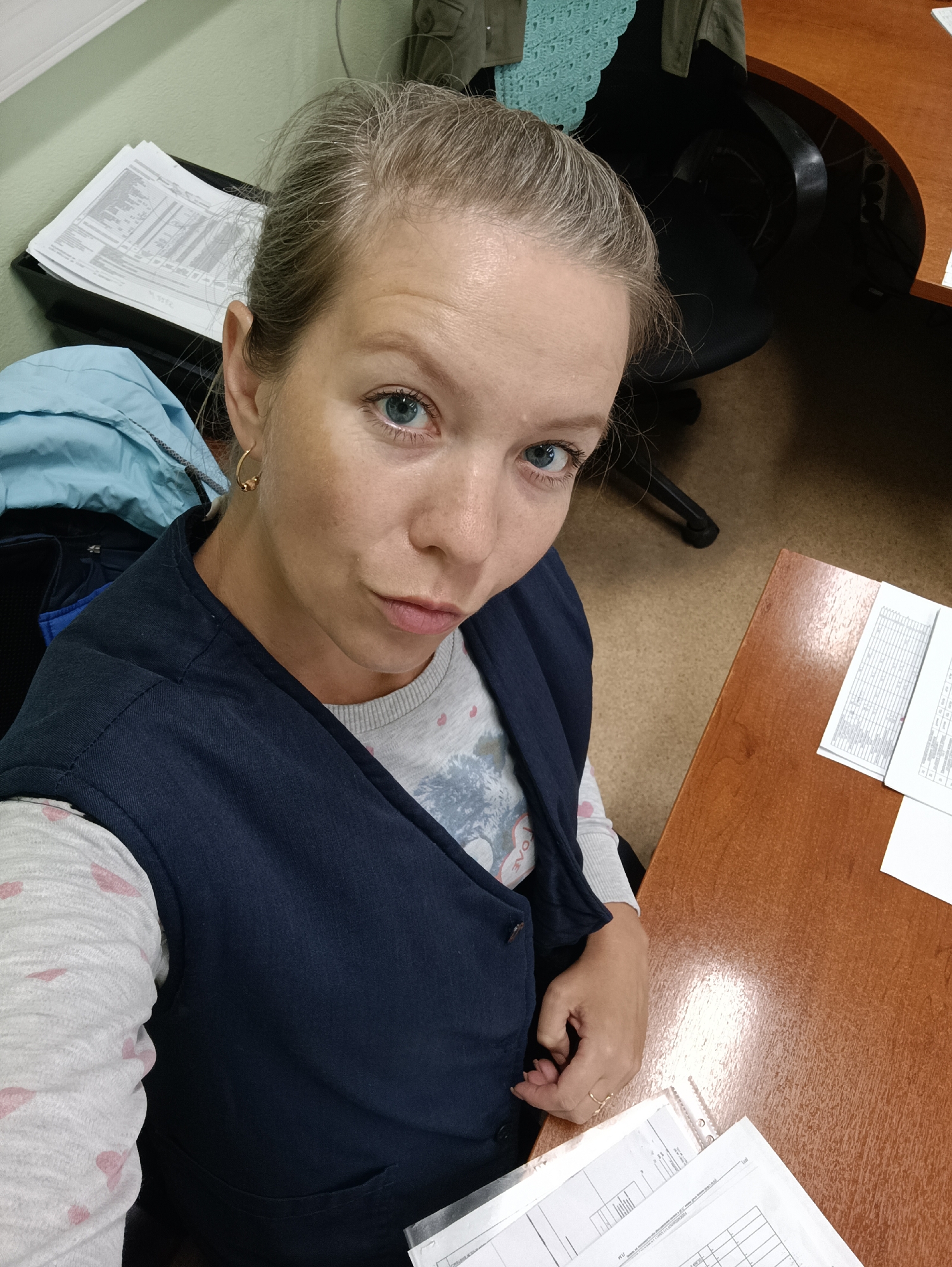 A woman with light skin and blue eyes is taking a selfie from an elevated angle. She has her hair pulled back and is wearing a dark blue vest over a light gray long-sleeved shirt. She has a serious expression with slightly pursed lips. She is wearing gold hoop earrings and a gold ring on her finger.
The setting appears to be an office. There are documents and papers on a wooden desk to her right and on a pile of papers on a black tray to her left. A blue jacket and a black office chair are also visible in the background.
There is no definitive information in the image that confirms the location as Chaikovsky, Russia.