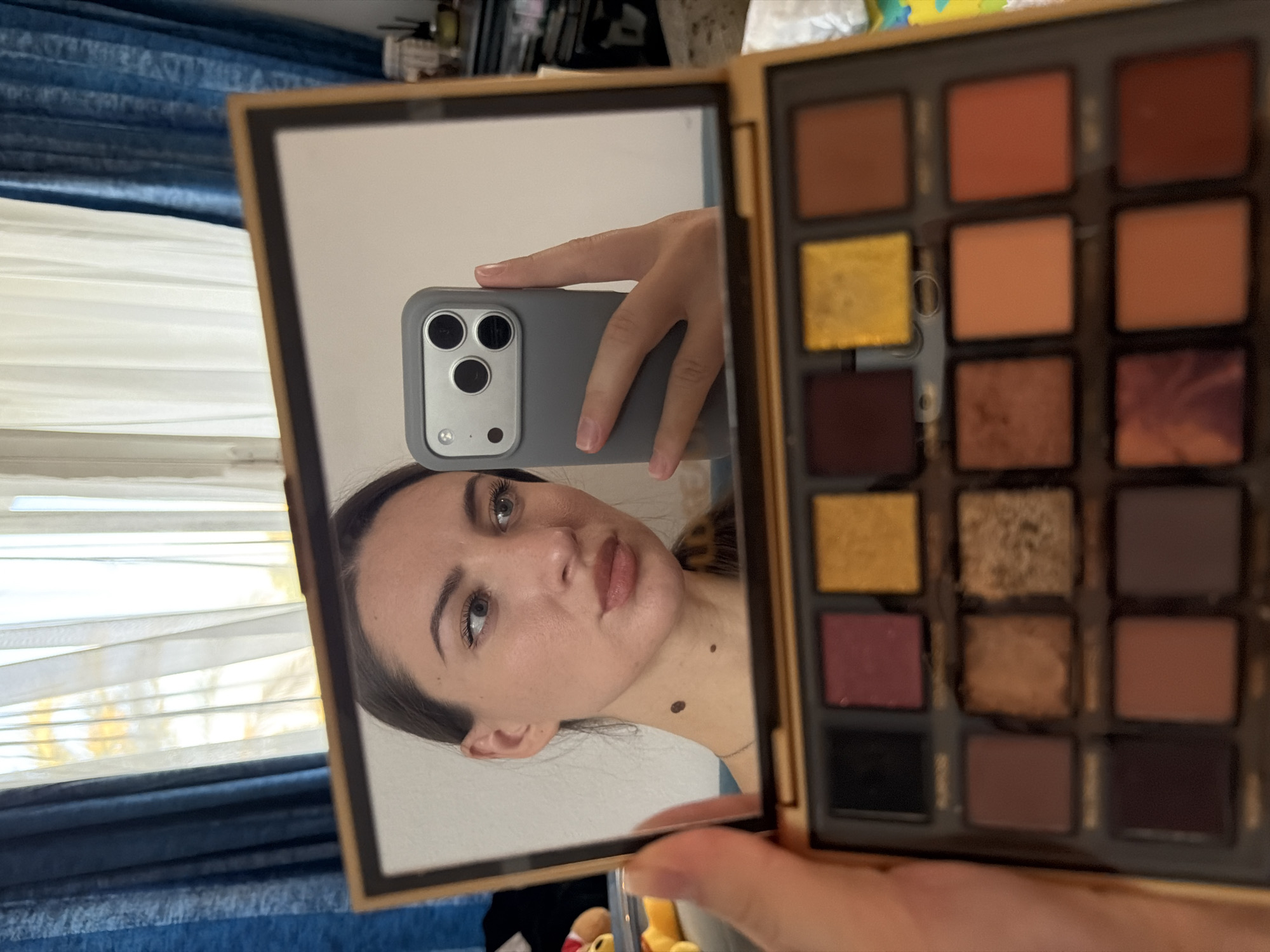 A woman is holding a makeup palette and taking a selfie in a mirror. The makeup palette is open, revealing a collection of eyeshadows in various shades of brown, red, and yellow. The woman is looking at the mirror, and her iPhone is visible in the reflection. She has light brown hair pulled back and is wearing subtle makeup. The background shows a window with sheer white curtains and blue drapes. The image is likely taken indoors, possibly in a residential setting in Sharm El Sheikh, Egypt, given the curtains and light.