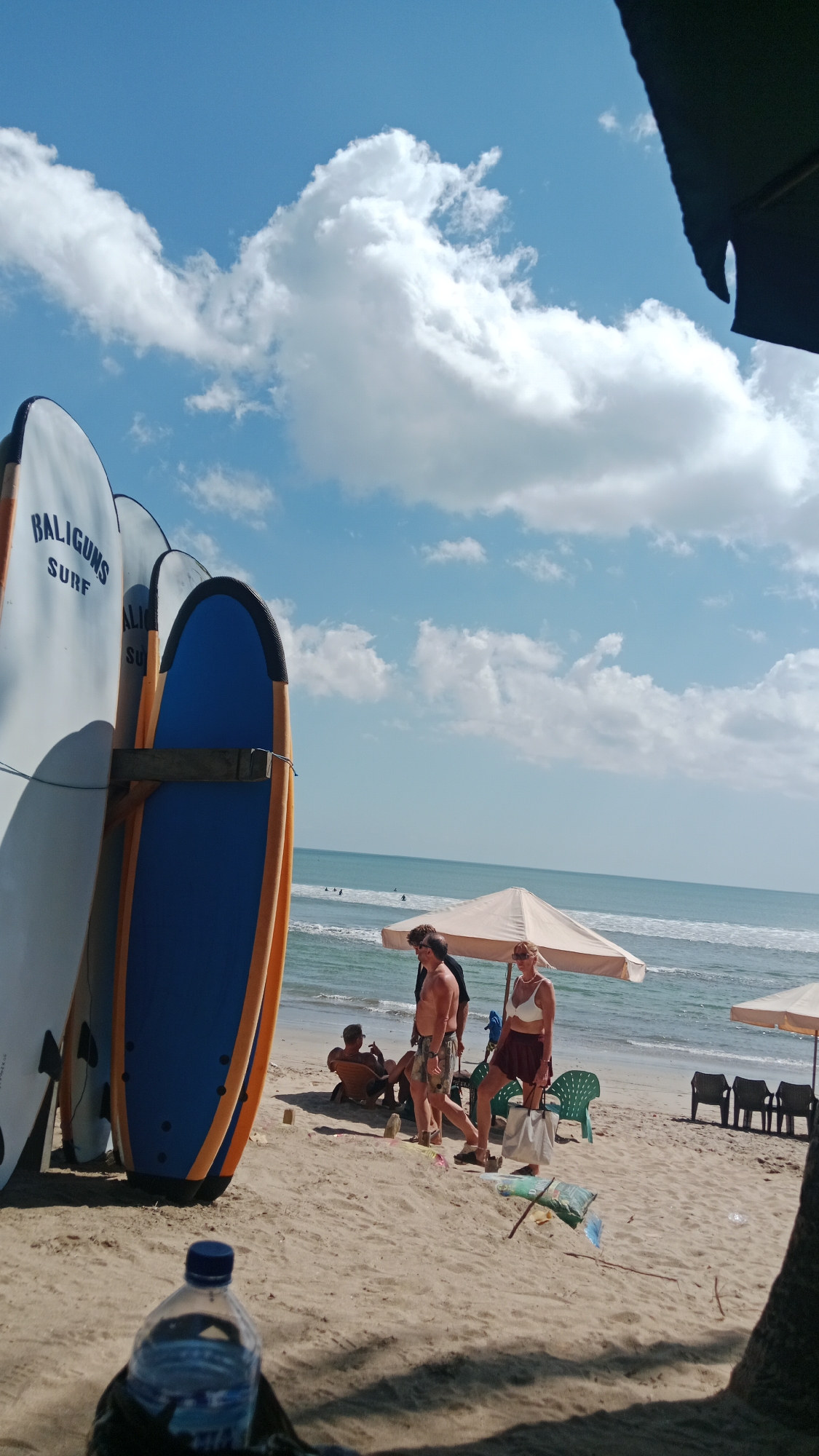 A sunny day at Kuta Beach in Bali, Indonesia, is depicted, with a clear blue sky dotted with white clouds. In the foreground, a partially visible water bottle sits on the sand. To the left, several surfboards are propped upright, one with "BALIGUNS SURF" written on it.
In the middle ground, a group of people are on the sandy beach. Two men and one woman are walking near a beach umbrella. One man, wearing patterned shorts, is walking away from the camera. Another man, in shorts, walks towards the camera. A woman in a white top and a maroon skirt is also walking. Another man is seated in a lounge chair further back.
The ocean stretches out to the horizon, with small waves breaking. Several surfers can be seen in the water. Beach umbrellas and chairs are set up on the sand, suggesting a typical beach scene in Kuta. The overall atmosphere is one of leisure and activity.