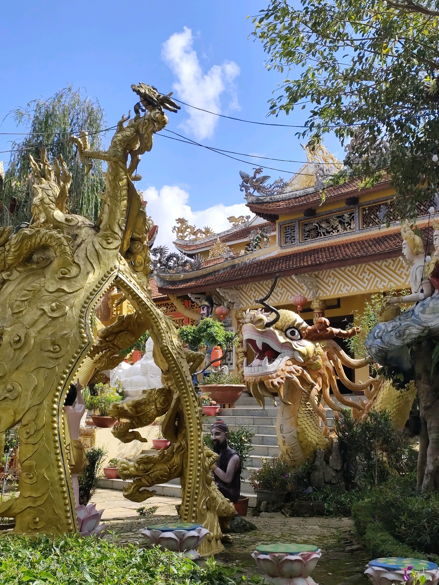The image depicts a scene outside a temple in Da Lat, Vietnam. Prominently featured are large, ornate golden dragon statues, one with its head and open mouth facing the viewer, and another coiled structure in the foreground. Behind the statues, a traditional temple building with a tiled roof and decorative elements is visible, with stairs leading up to its entrance. The surrounding area includes lush green foliage, potted plants, and what appear to be lotus-shaped decorative elements on the ground. The sky is blue with scattered clouds, and sunlight illuminates the scene. Several figures, including what looks like a statue of a robed man with a beard, are also present within the temple grounds. Electrical wires are strung across the upper portion of the image.