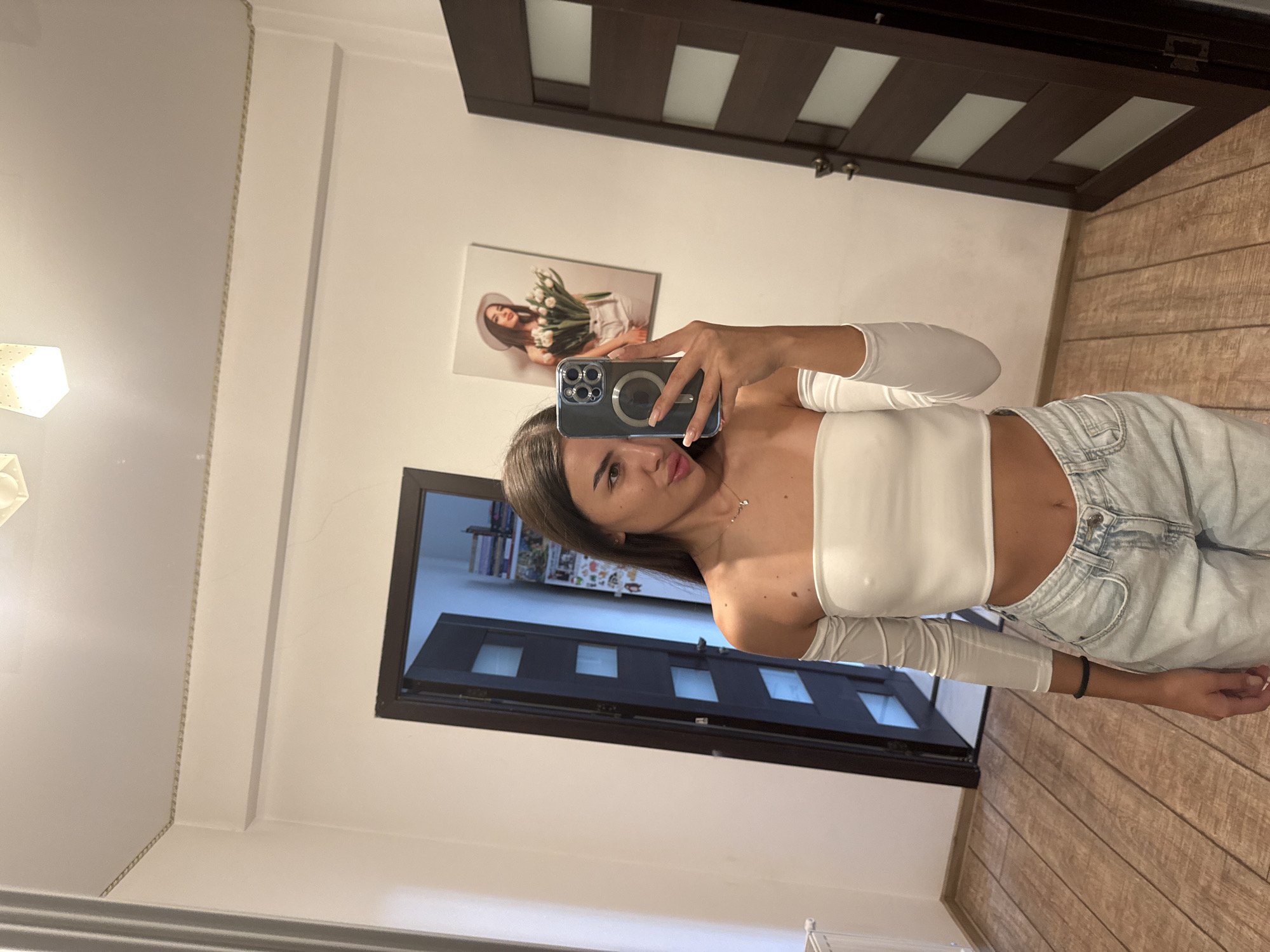 A young woman with long brown hair is taking a selfie in a room with wooden floors and white walls. She is wearing a white off-the-shoulder crop top and light-wash jeans. In the background, there is an open door leading into another room, a radiator on the left, and a framed picture on the wall to the right of the open door. The framed picture appears to be of a woman wearing a white hat and holding a bouquet of tulips. The room's ceiling has recessed lighting. The scene is likely taking place indoors, possibly in a residential setting, in the city of Chișinău, Moldova.