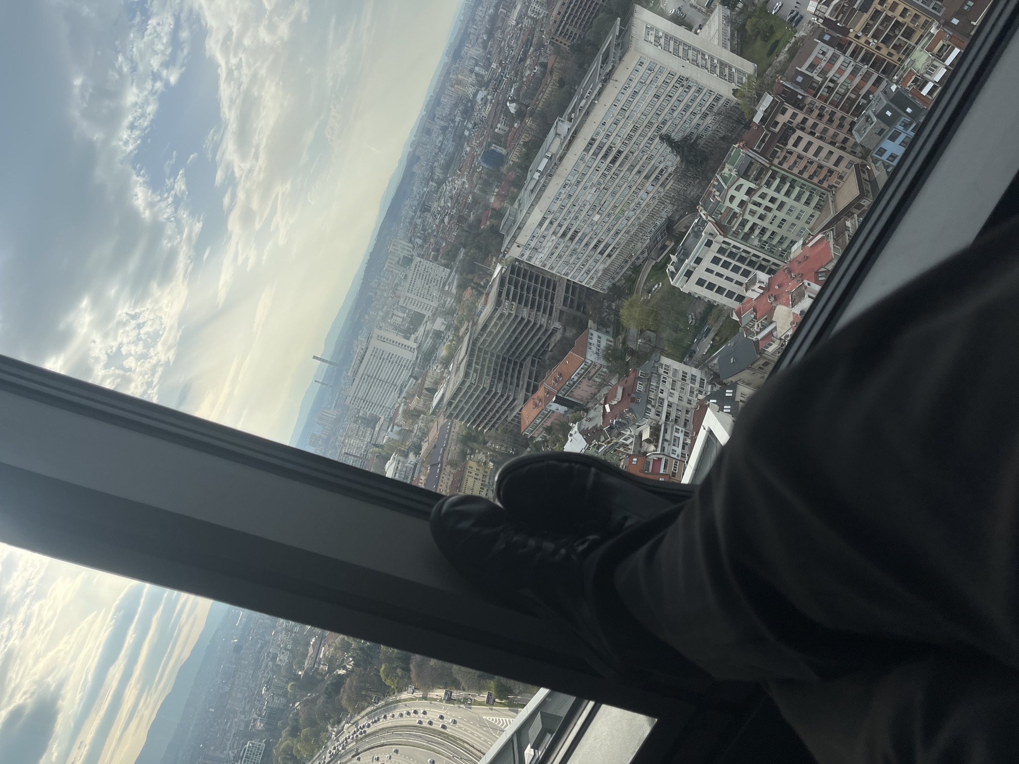 A medium shot shows a pair of legs and feet in black shoes resting on a window sill, with a cityscape visible outside. The photograph appears to be taken from a high vantage point, likely an upper floor of a building. The cityscape shows a dense urban area with a mix of high-rise buildings and lower structures, indicative of a large city. There are many buildings in the background. Based on the architecture and landscape, the city is identified as Sofia, Bulgaria. The sky is visible in the upper portion of the frame, showing scattered clouds and sunlight. The image orientation is not straight, with the view skewed to an angle.