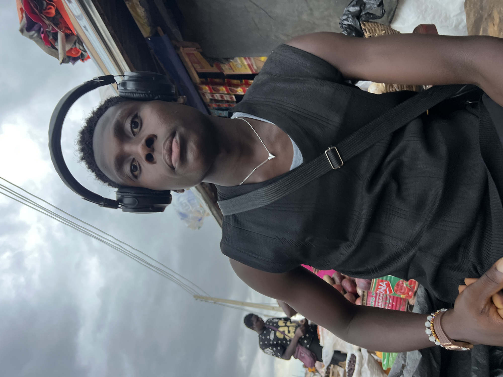 A young Black male with dark skin, short curly hair, and headphones on his head is looking directly at the camera. He is wearing a black sleeveless shirt over a gray t-shirt, a silver necklace with a heart pendant, and a brown watch with a band on his left wrist. He has a black crossbody bag with a silver buckle. The background appears to be an outdoor market in Asaba, Nigeria, with various stalls and goods visible, including canned goods and produce. The sky is overcast. Another person in a black shirt with white polka dots and a red bag is visible in the background. The overall scene suggests a bustling market environment.