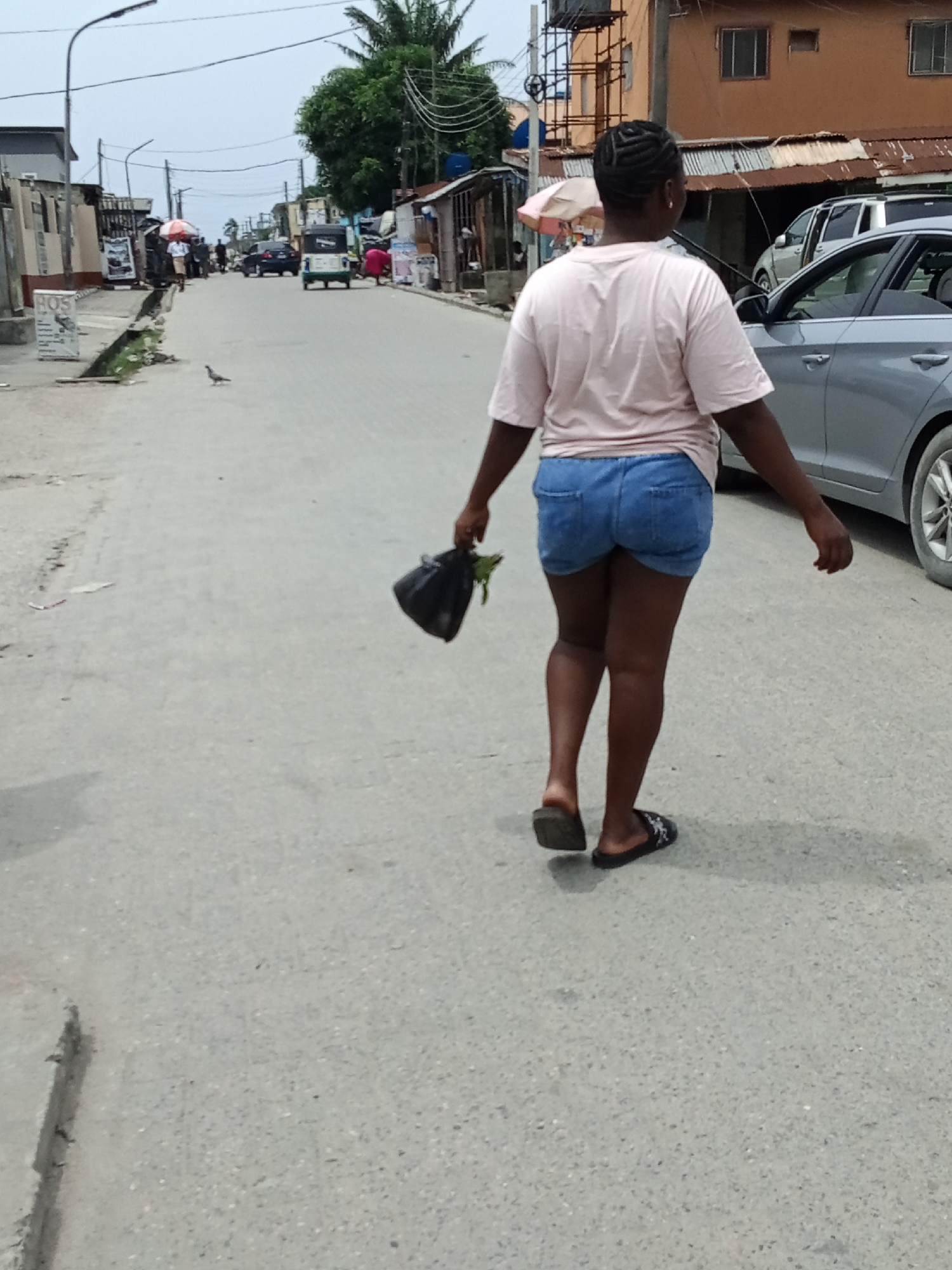 A street scene shows a woman walking away from the viewer down a grey asphalt road. She is carrying a black plastic bag. The woman is wearing a light pink t-shirt, blue denim shorts, and black sandals. To the right of the woman is a silver car. On the left side of the road, there is a small bird. In the background, there are shops and pedestrians.