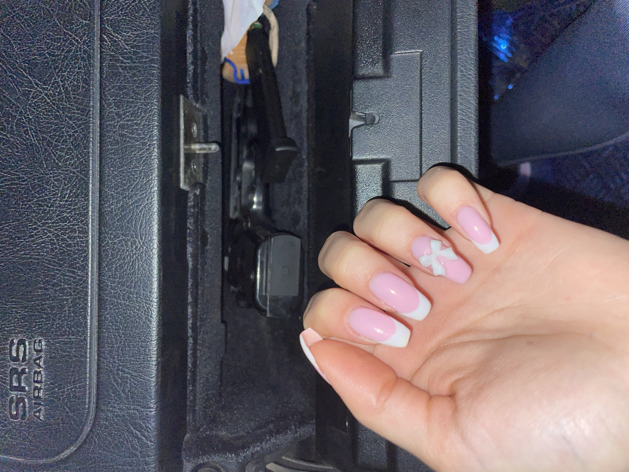 A close-up captures a hand with manicured nails against the interior of a car. The nails are pastel pink with white tips, and a small white bow adorns one nail. The car's interior features a black, textured plastic component labeled "SRS AIRBAG," and a storage compartment appears partially open, revealing mechanical elements within. The location is not determinable from the image.