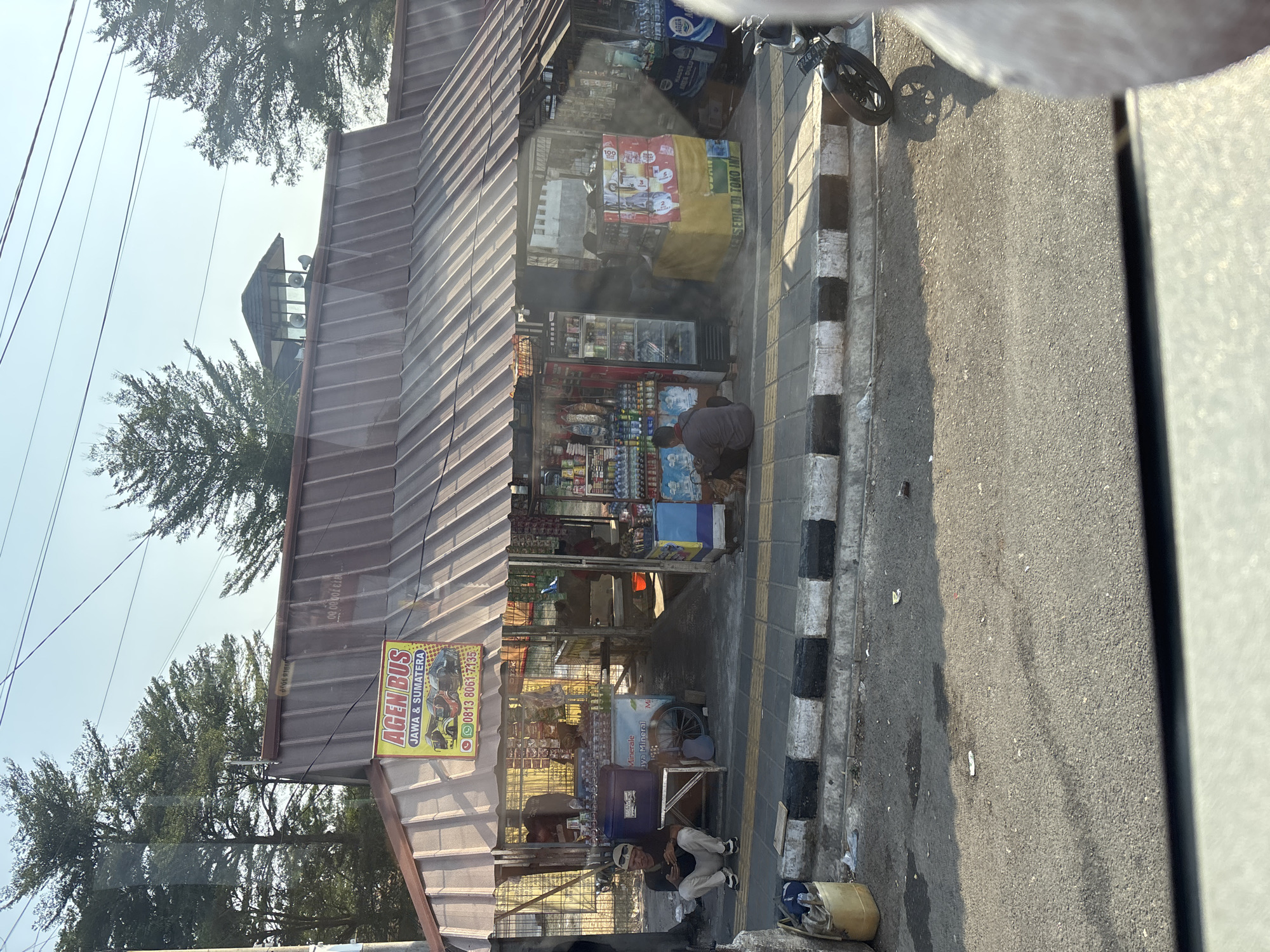 The image depicts a bus ticket and convenience store in Cikampek, Indonesia, likely on a roadside. The storefront has a corrugated metal roof and displays an "AGEN BUS JAWA & SUMATERA" sign. Inside, shelves are stocked with various goods, including bottled drinks and snacks. A refrigerator unit is visible on the left side.
Two men are present outside the store. One man, wearing a white cap, is seated on the curb in front of the store, looking at his phone. He is wearing a dark shirt and light-colored pants. Another man is seated on the ground inside the covered area of the store, also appearing to be looking down.
In front of the store, a sidewalk with a black and white striped border runs along the street. A yellow plastic container and some discarded items are on the ground next to the sidewalk. A motorcycle is parked on the street to the right of the scene, casting a shadow on the asphalt. The sky is clear and blue, suggesting it is daytime. The overall environment suggests a commercial area with a focus on transportation services and local retail.