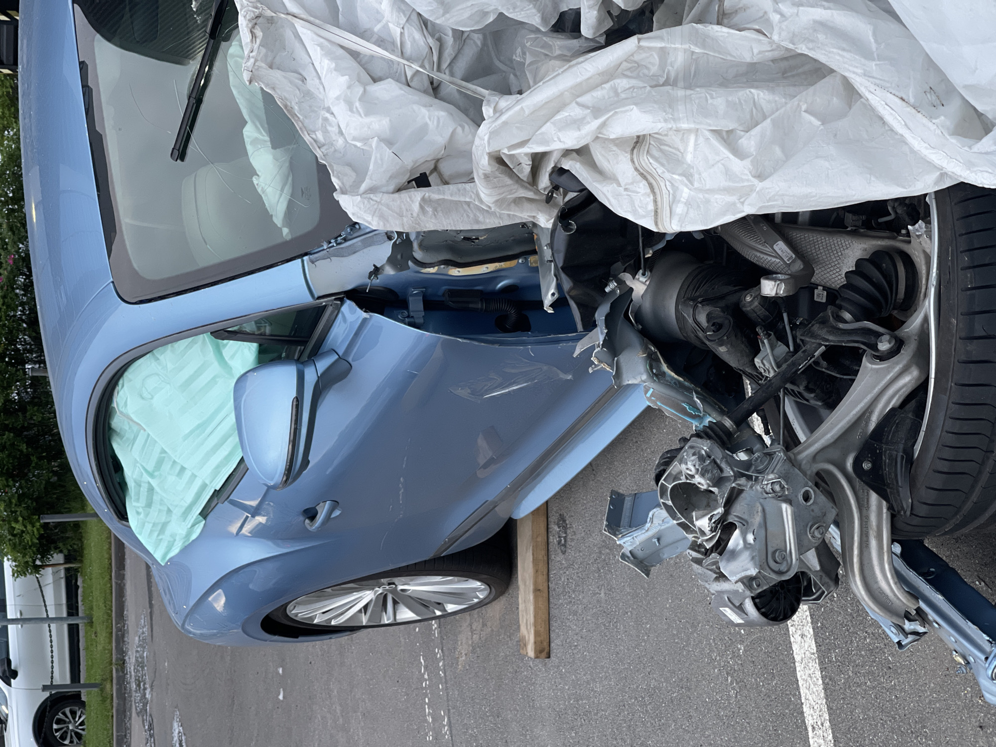A blue car sits damaged in an asphalt parking lot. The driver's side front of the car is heavily damaged, with the fender and hood detached, revealing a complex assembly of mechanical parts. The airbags have deployed, covering the interior of the car and obscuring the windshield. A white tarp is draped over some of the damaged components. The car's front tire is visible, resting on a wooden block. In the background, a white SUV is parked, and a low green fence with bushes behind it separates the parking lot from a grassy area. The ground shows some scattered debris.