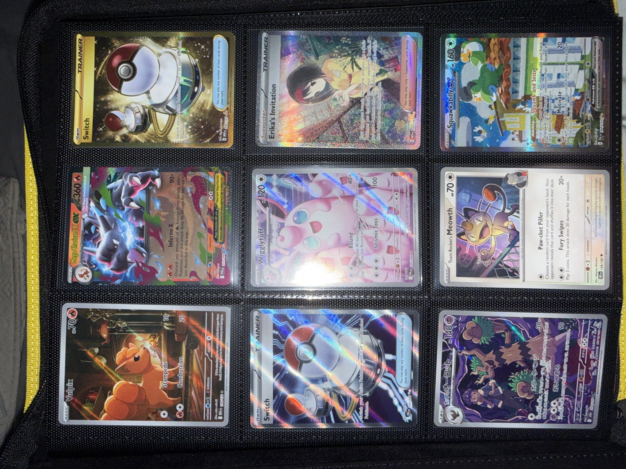 The image shows a collection of Pokémon trading cards displayed in a black binder with yellow trim. The binder is laid flat, and the cards are organized into rows within plastic sleeves. The cards depicted are:
Row 1:
* A Vulpix card on the left.
* A Mega Charizard EX card in the center.
* A "Switch" Trainer card with a gold border on the right.
Row 2:
* A "Switch" Trainer card with a holographic, non-gold border on the left.
* A Wigglytuff card in the center.
* An "Erika's Invitation" Supporter card with a holographic border on the right.
Row 3:
* A Japanese Pokémon card with dark purple and black imagery, featuring a tree-like creature, on the left.
* A "Team Rocket's Meowth" card in the center.
* A "Squawkabilly EX" card with a holographic border on the right.
The lighting appears to be artificial, casting a slight sheen on the holographic cards. The background is a dark, textured material, consistent with the inside of a binder. The specific location within a city or any environmental details beyond the binder itself are not ascertainable.
