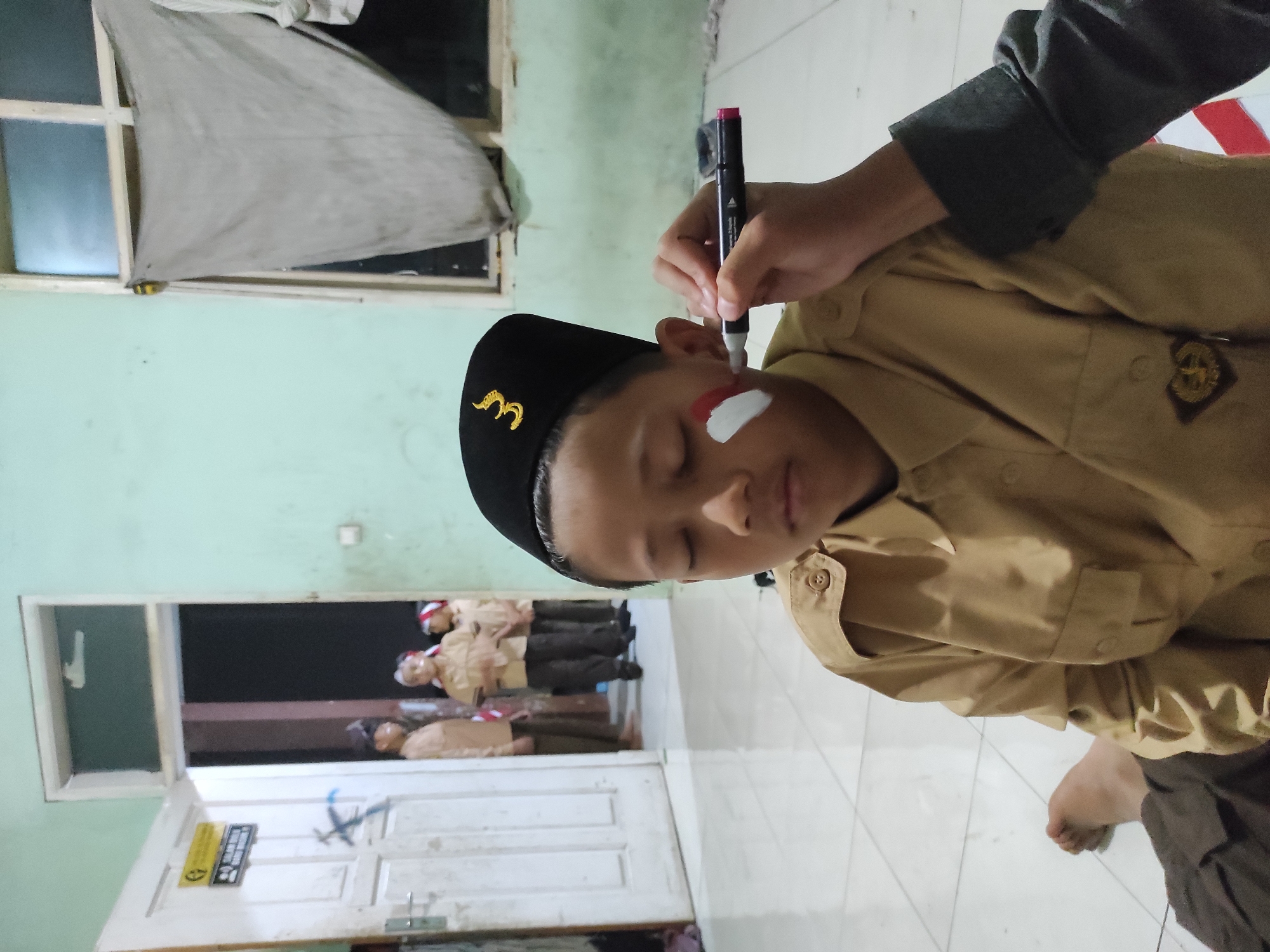 A young Indonesian boy, wearing a tan uniform and a black cap with a gold emblem, is seated on a tiled floor. He has his eyes closed while someone, whose arm and hand are visible in the foreground, is painting red and white stripes on his cheek with a marker. The stripes appear to be the colors of the Indonesian flag. In the background, through an open doorway, other children in similar uniforms are visible. The environment appears to be indoors, possibly a school or community building, with light green walls and a window with a light-colored fabric draped over it. The overall impression is of preparation for a patriotic event.