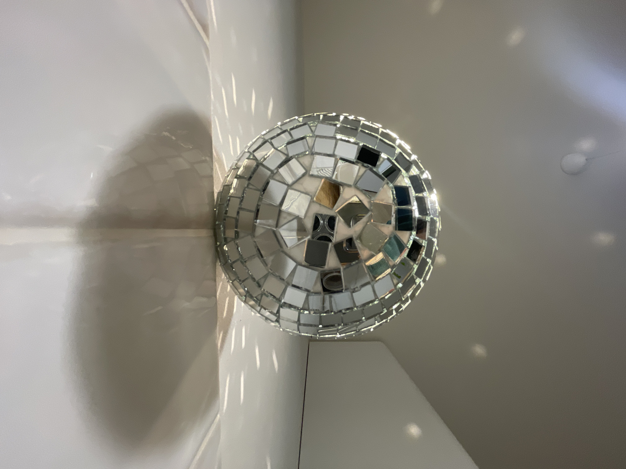 A disco ball is mounted in a corner of a room. The walls are white, and light reflects off the mirrored tiles of the disco ball, creating scattered patterns on the walls and ceiling.