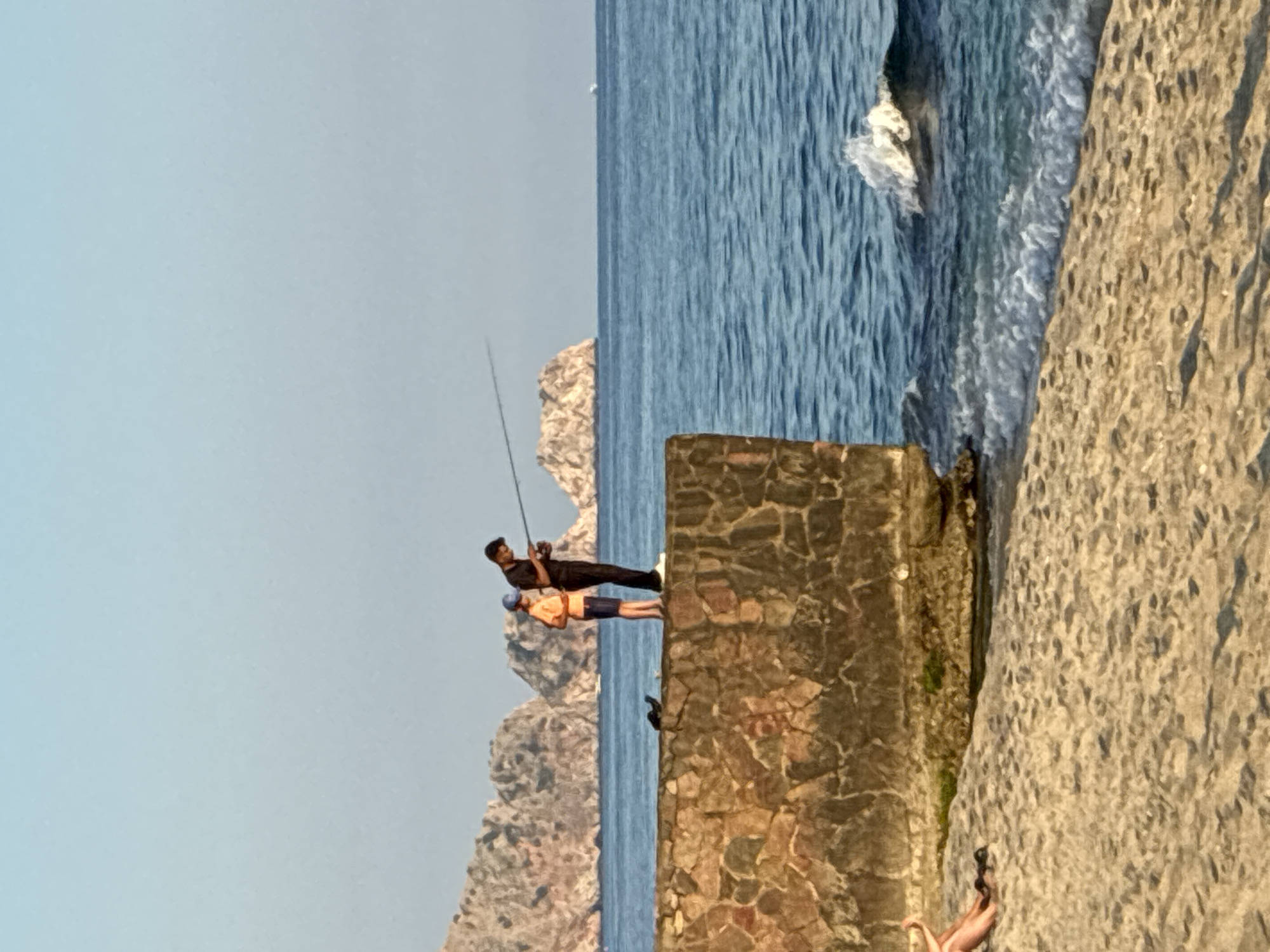 The image depicts a scenic view of the Mediterranean coast in Alanya, Turkey. In the foreground, a sandy beach is visible with the gentle waves of the sea lapping at the shore. A stone wall, constructed from irregular, multi-colored stones, forms a prominent feature, extending into the water. Two figures are standing on top of this wall, engaged in fishing. One is a young person wearing an orange shirt and blue shorts, holding a fishing rod. Beside them, an adult, dressed in a black shirt and pants, is also fishing. In the background, the distinctive rocky hills and cliffs of Alanya are visible, framing the expansive blue sea. A small boat can be seen in the distance, adding to the maritime atmosphere. The sky is clear and a pale blue, indicative of a sunny day.