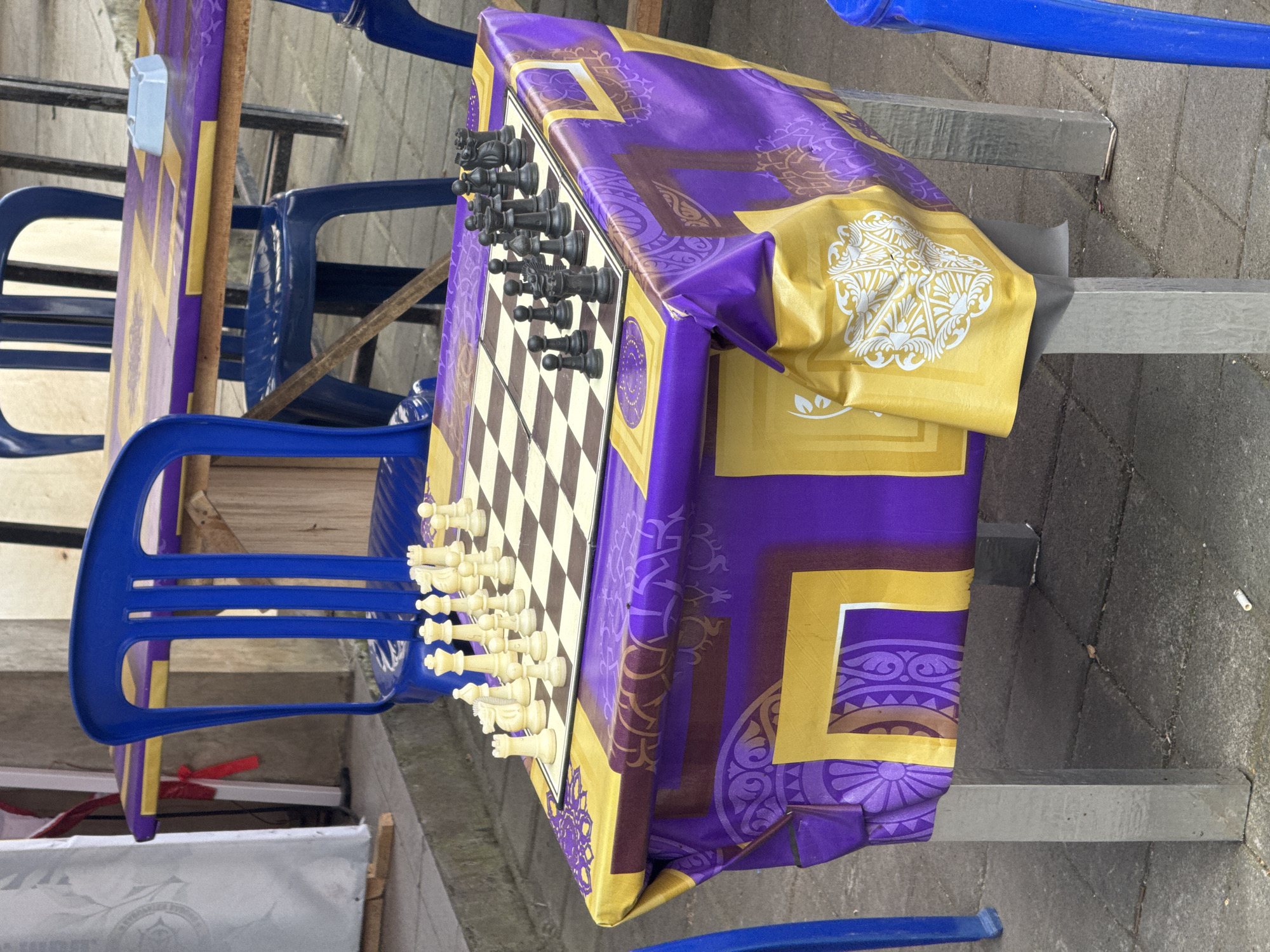In Kuta Selatan, Indonesia, a chessboard sits on a table covered with a purple and yellow patterned cloth. The chess set is partially arranged, with white pieces on one side and black pieces on the other. Several blue plastic chairs are visible around the table. The table is supported by metal legs and is situated outdoors on a paved surface. A discarded cigarette butt lies on the ground near the table. The background includes a white curtain or fabric structure and other tables and chairs.