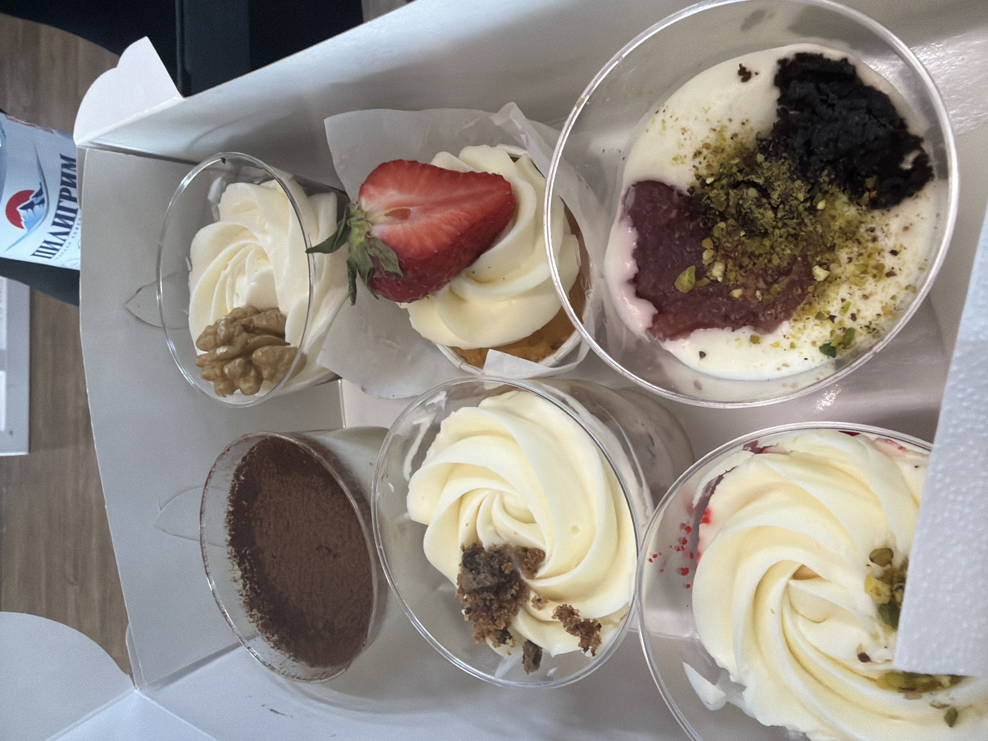 An overhead shot captures a white cardboard box filled with an assortment of six individual desserts in clear plastic cups. The box appears to be open, showcasing the sweet treats within.
On the top row, from left to right:
1. A clear cup with swirled white frosting, garnished with whole walnuts.
2. A cupcake in a white paper liner, topped with white frosting and a fresh, halved strawberry.
3. A clear cup holding a creamy white dessert, featuring distinct toppings of dark crumbs (possibly chocolate or Oreo), a red fruit compote or jam, and scattered green crushed pistachios.
On the bottom row, from left to right:
1. A clear cup filled with a light brown powdery substance, likely cocoa, suggesting a dessert like tiramisu.
2. A clear cup with swirled white frosting, adorned with light brown crumbs.
3. A clear cup with swirled white frosting, sprinkled with small red specks and a few green crushed nuts, possibly pistachios.
To the left of the box, partially visible, is a blue and white bottle with the Cyrillic text "ПИЛИГРИМ" (PILIGRIM) printed on its label, indicating it is likely a bottle of Pilgrim mineral water, consistent with the Chaikovsky, Russia location context. The setting appears to be indoors, under a bright, even light, highlighting the details of each dessert. The arrangement suggests the desserts are either packaged for sale, catering, or a special occasion.