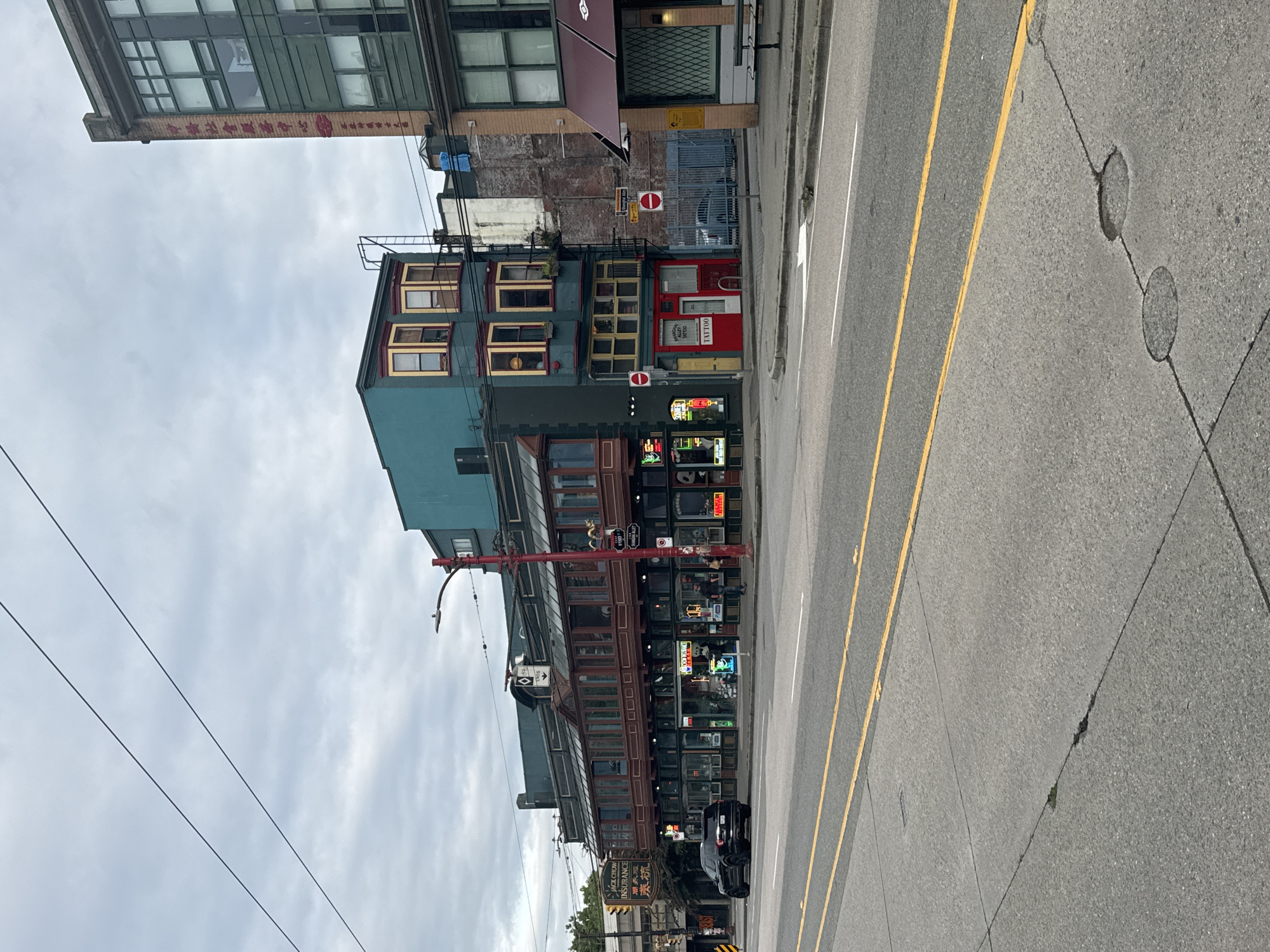 The image depicts a street scene in Chinatown, Vancouver, Canada, on an overcast day. The foreground features an asphalt road with double yellow lines and some cracks. In the midground, a row of buildings with distinct architecture lines the street. The buildings have multiple stories, with the lower levels featuring storefronts with neon signs, including "Jack Chow Insurance" and what appears to be "Sushi". The upper levels have a more traditional East Asian aesthetic with dark red wooden balconies and bay windows. One building is painted a striking teal color. Several people are visible on the sidewalk, appearing to be pedestrians. Red poles, possibly for streetlights or decorative elements, are prominent. A black SUV is parked on the side of the street. The background shows more buildings, some with visible signage, and a cloudy sky.