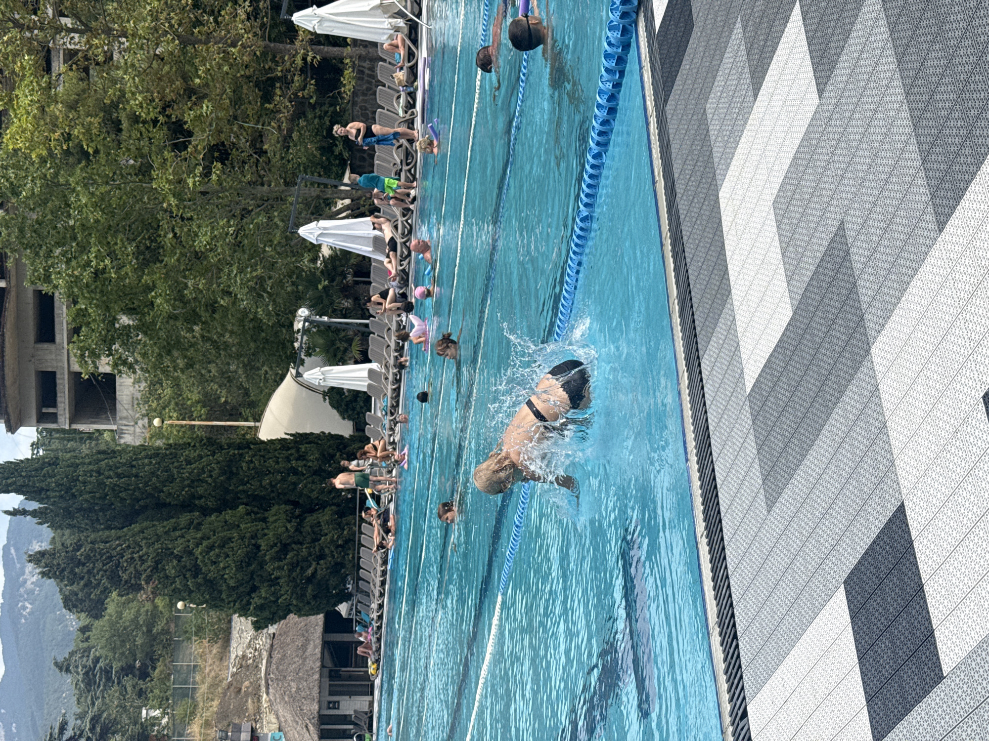 The image depicts a crowded outdoor swimming pool area, likely at a resort or aquatic center. The pool itself is large and divided into lanes by blue lane ropes. Several people are swimming in the pool, including a woman in the foreground who is mid-splash, seemingly diving or jumping into the water. Other individuals are seen swimming, some with pool noodles, while a few are lounging on chairs on the poolside. In the background, there are numerous lounge chairs, some occupied by sunbathers, and large white umbrellas providing shade. A building with multiple stories and an unfinished facade is visible behind the pool area, along with dense green trees and hills in the distance. The poolside is paved with a patterned tiling in shades of grey and white. This scene is characteristic of recreational facilities found in a city like Yalta, Ukraine, known for its coastal resorts and summertime attractions.
