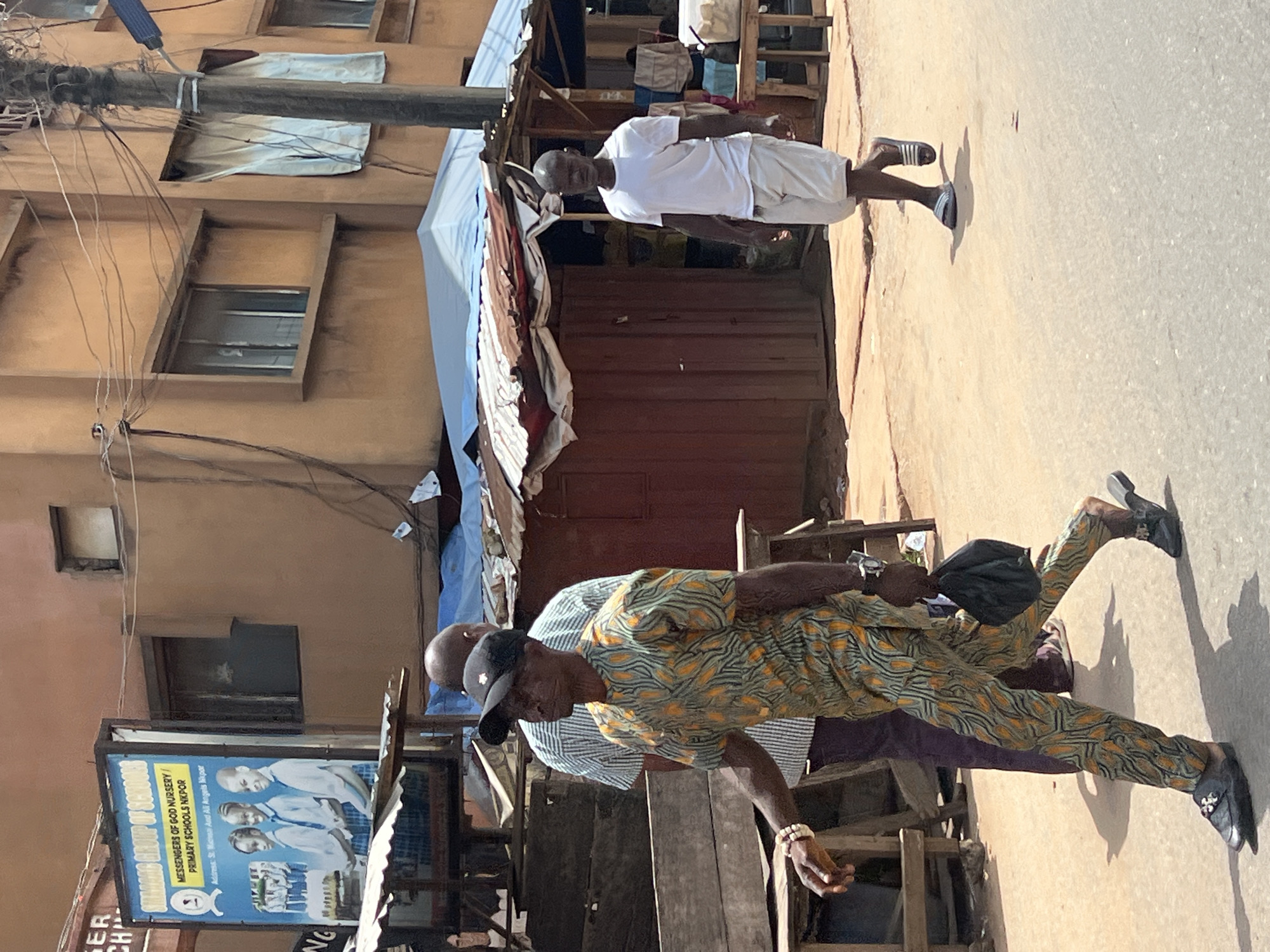 A street scene in Nkpor, Nigeria, features a brown building with multiple windows and a utility pole with many wires. A blue billboard for "SHAGAMA GROUP OF SCHOOLS" displays images of three children in school uniforms and text. In the foreground, three men are visible. The man on the left wears a patterned green and orange tunic, dark pants, a black baseball cap, and a bracelet. He is walking and carrying a black bag. Behind him, another man in a striped shirt and dark pants is also walking. To the right, a man in a white t-shirt and white shorts walks away from the camera. The ground is paved with asphalt and a lighter-colored concrete or dirt pathway. Several makeshift structures with corrugated metal roofs are present, one with a red wooden front. The scene appears to be an outdoor market or commercial area.