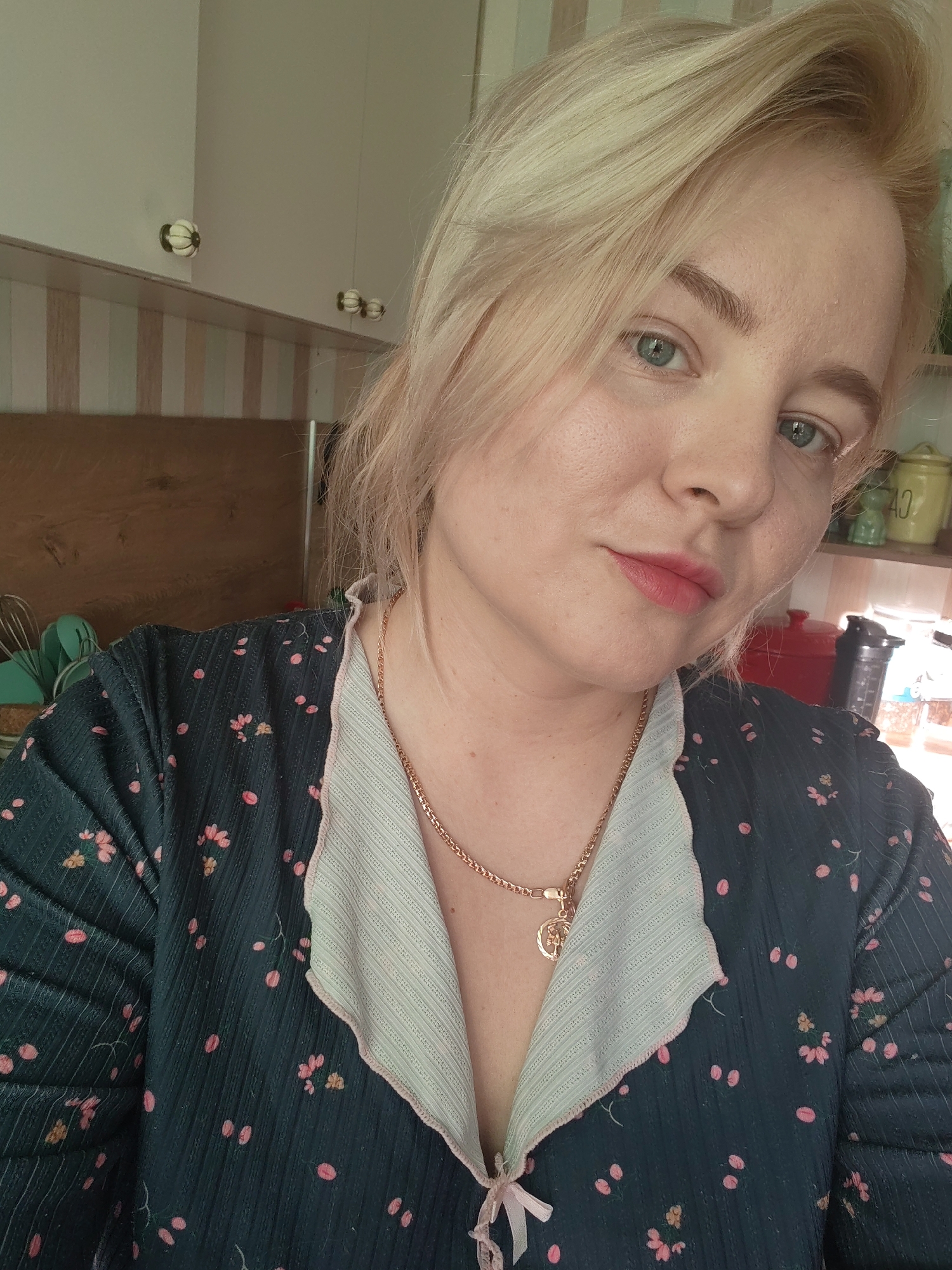A young woman with fair skin, light blonde hair styled loosely to the side, and blue eyes is posing for a selfie in what appears to be an indoor setting, possibly a kitchen. She is wearing a dark green patterned top with pink floral designs and a light green collar that is tied with a pink ribbon. A gold necklace is visible around her neck. The background shows kitchen cabinets in white and striped patterns. Kitchen utensils and jars are also visible on a shelf behind her. The location is likely in Izhevsk, Russia, based on the information requested.
