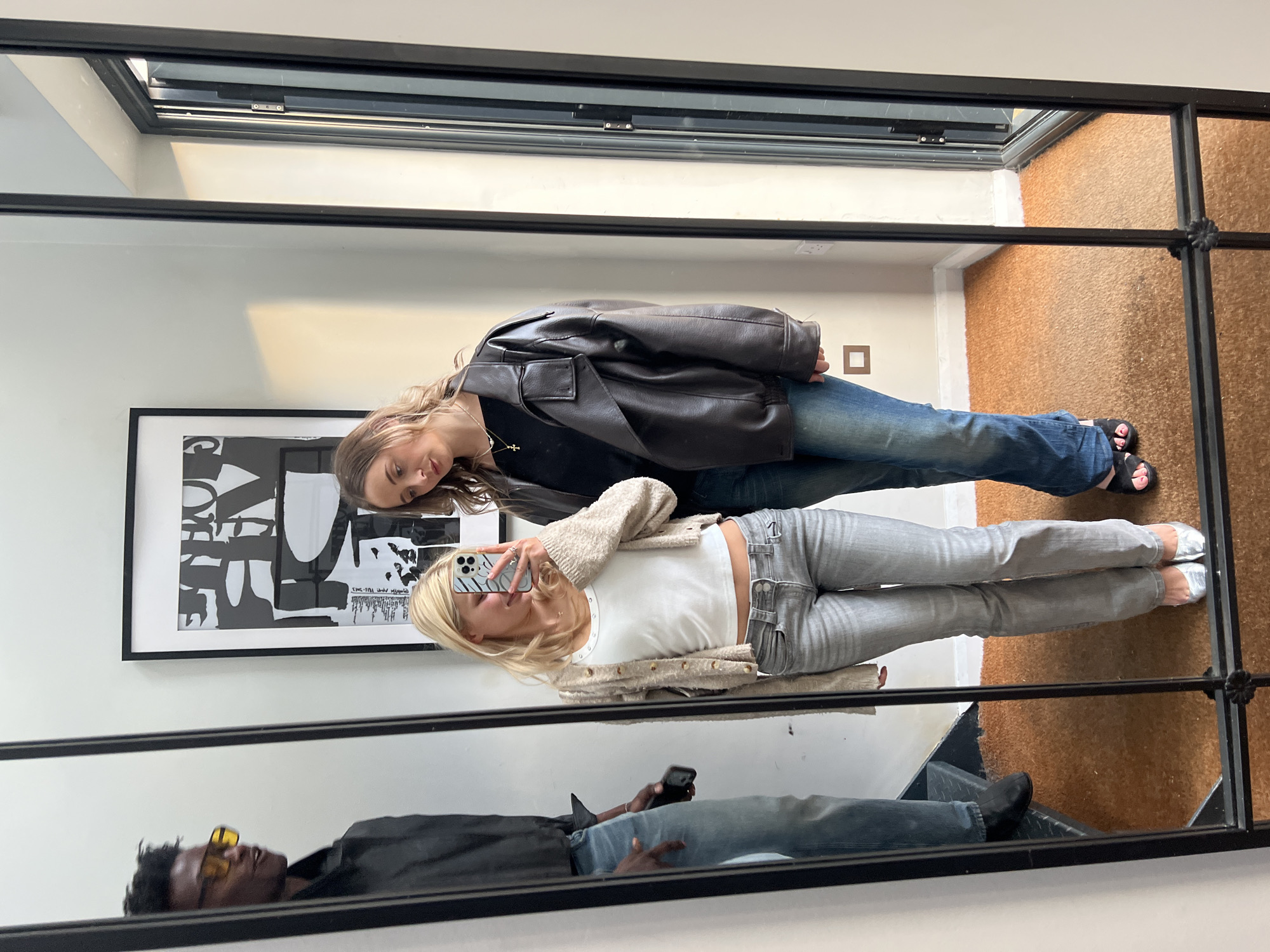 Two women are posing for a photo in front of a large, black-framed mirror. The woman at the top is wearing a black top, blue jeans, and a brown leather jacket. The woman below is wearing a white top, grey jeans, and a beige cardigan. The blonde is holding a phone in front of her face, presumably taking the photo. In the reflection, a man is visible. He is wearing sunglasses, a black shirt, and jeans.
The scene appears to be indoors, possibly in an apartment or shop. The floor is a brown carpet or mat. A framed print is visible on the wall behind the women. Above is a window.
Without additional information, pinpointing the precise location in London is impossible.