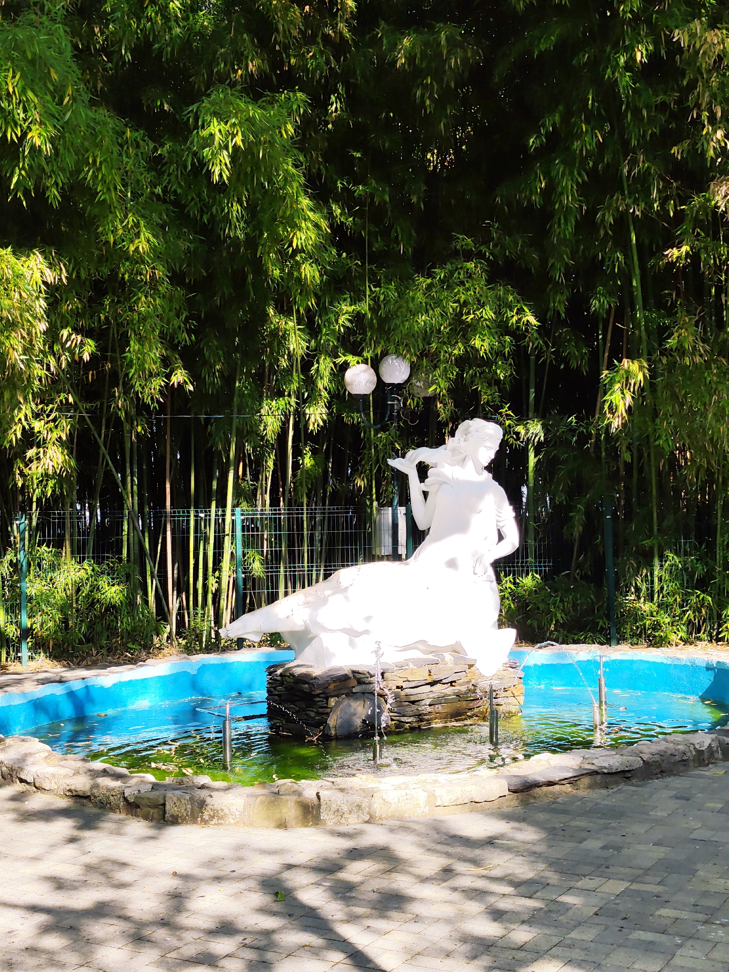 This image captures a serene outdoor scene featuring a white sculpture of a woman in a flowing dress, standing in a vibrant blue fountain. The sculpture is positioned on a dark, rocky base within the water, which has a greenish tint and several small fountains. The fountain is surrounded by a rustic stone border and sits on a paved area bathed in dappled sunlight, suggesting it's daytime with pleasant weather.
Behind the fountain, a dense grove of tall green bamboo trees creates a lush, natural backdrop. A metal fence separates the bamboo from the fountain area. Two white, spherical lanterns hang from a post near the bamboo, adding a decorative touch. The overall atmosphere is peaceful and picturesque, evoking a sense of tranquility. The location is identified as Sochi, Russia. No text is visible in the image.
