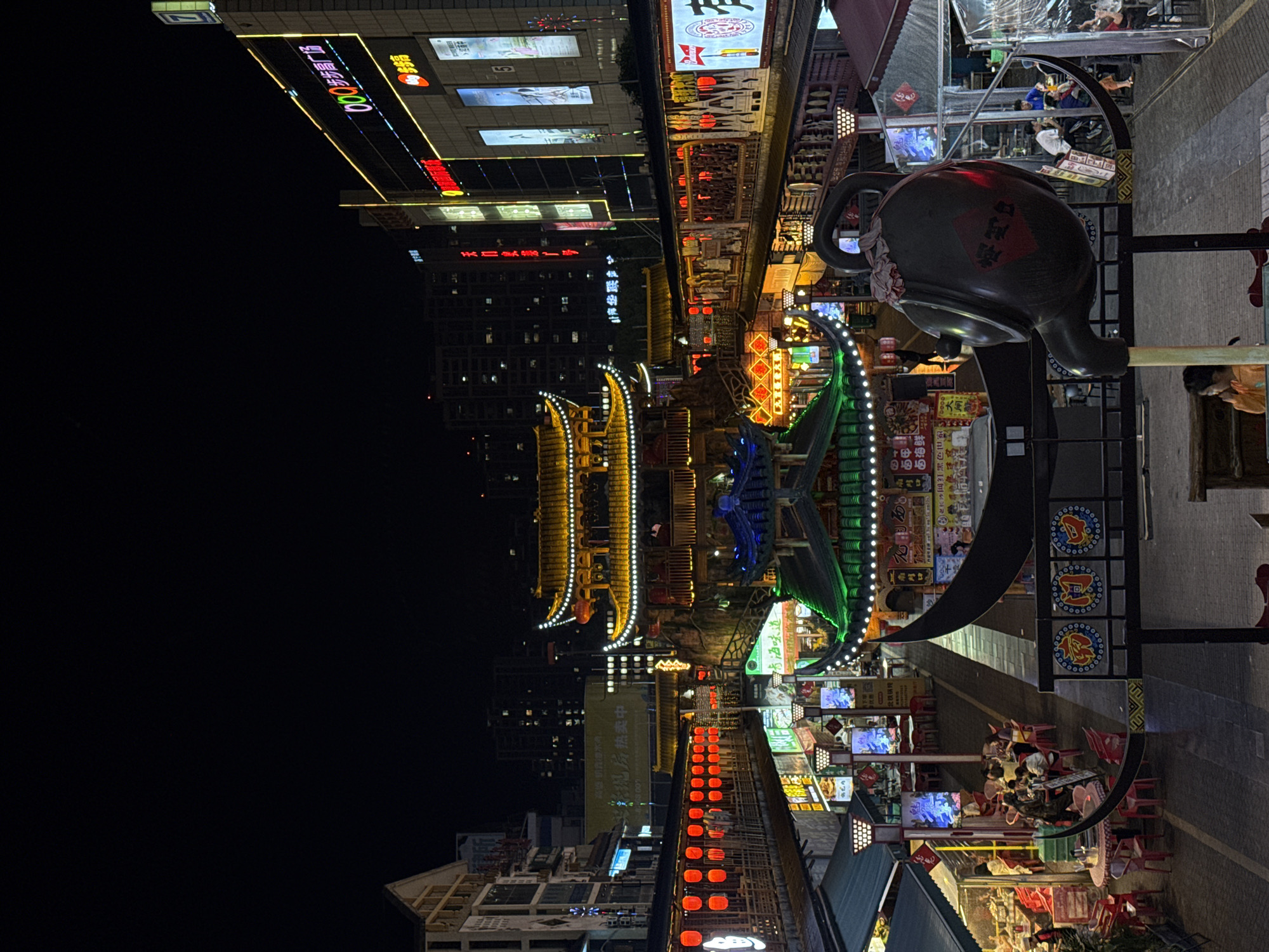 The image depicts a vibrant night scene on a commercial street in Zhangjiajie, China. The setting is illuminated by numerous artificial lights from shops, signs, and decorative elements, including traditional lanterns and illuminated architectural features.
In the foreground, a large, dark, decorative teapot sculpture with Chinese characters on its side stands prominently. To the right, people are seated at outdoor tables with red chairs, dining or socializing under ambient lighting. Various shops and restaurants line both sides of the street, many featuring illuminated signs and traditional Chinese architectural details, such as ornate roofs and decorative structures. A large building in the background on the left is also lit up with signage.
The atmosphere is lively and bustling, typical of a popular night market or pedestrian street. The extensive use of lighting highlights the cultural aesthetic of the area.
Visible text includes: "新现房热卖中" (New existing homes hot sale), "青海味道" (Qinghai flavor), "北铁锅烧" (Northern Iron Pot Roast), "南門口" (South Gate entrance), "步步高广场" (Bubugao Plaza), and "红黑色世家" (Red Black Family).