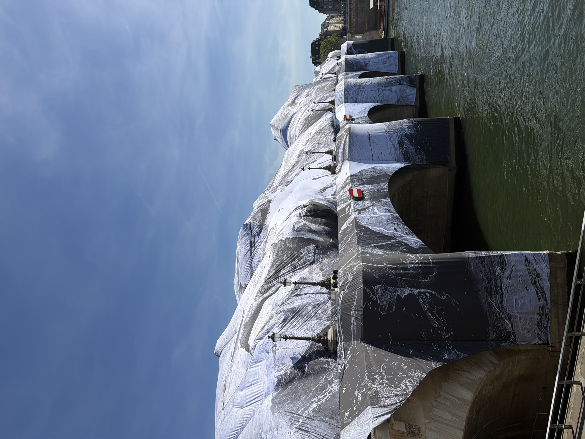 The image shows a bridge in Paris, France, wrapped in a silvery-white fabric with patterns of darker grey that resemble marble or flowing water. The bridge spans a body of water, its surface a slightly murky green. Parts of buildings are visible behind the bridge. The sky is pale blue with few clouds. There are black lampposts on the bridge.