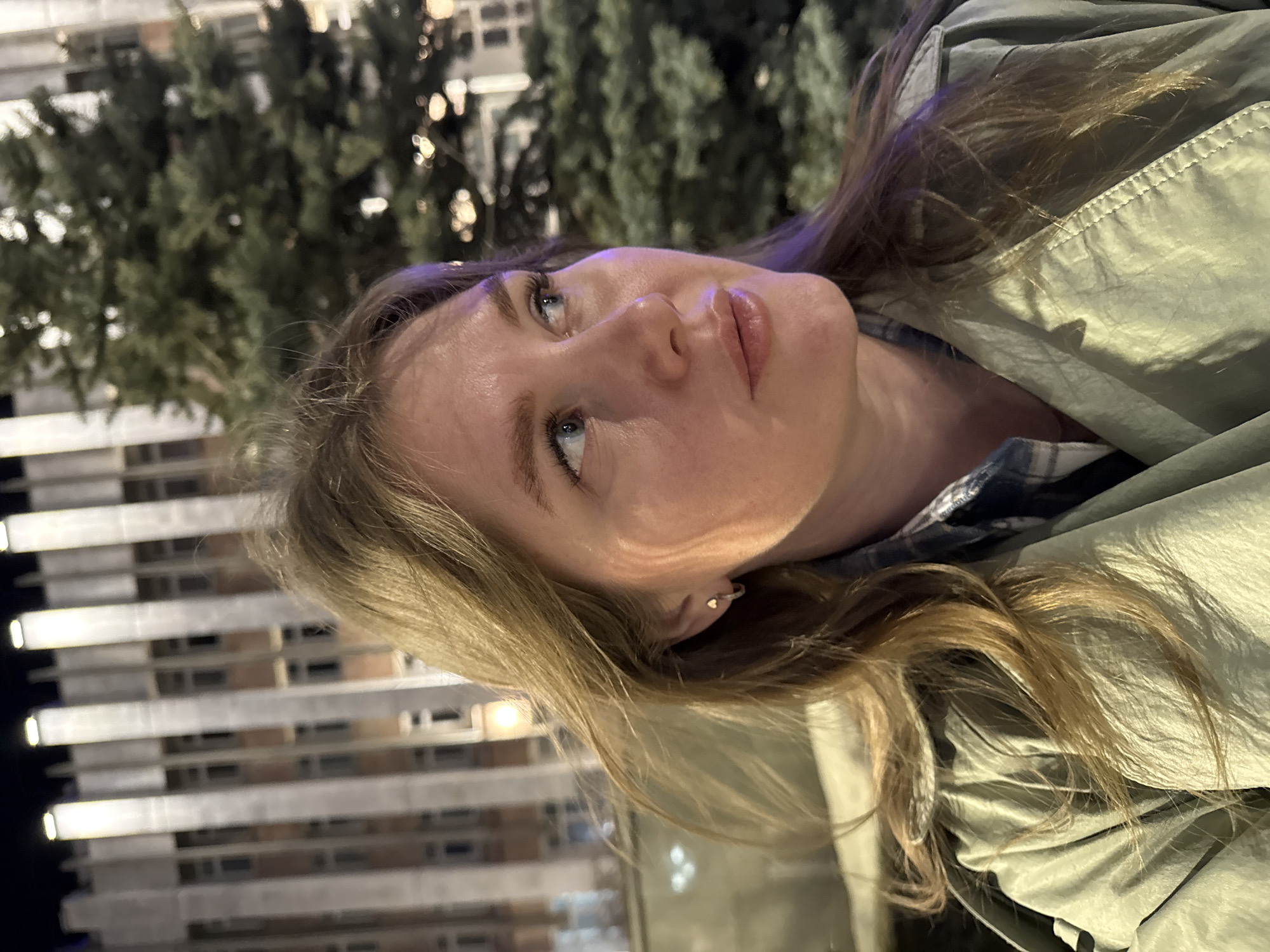 A photograph depicts a woman with light brown hair and fair skin. She is wearing a light green jacket over a plaid shirt. The background reveals urban elements. In the background are buildings with illuminated windows and evergreen trees. This scene is from Perm, Russia.