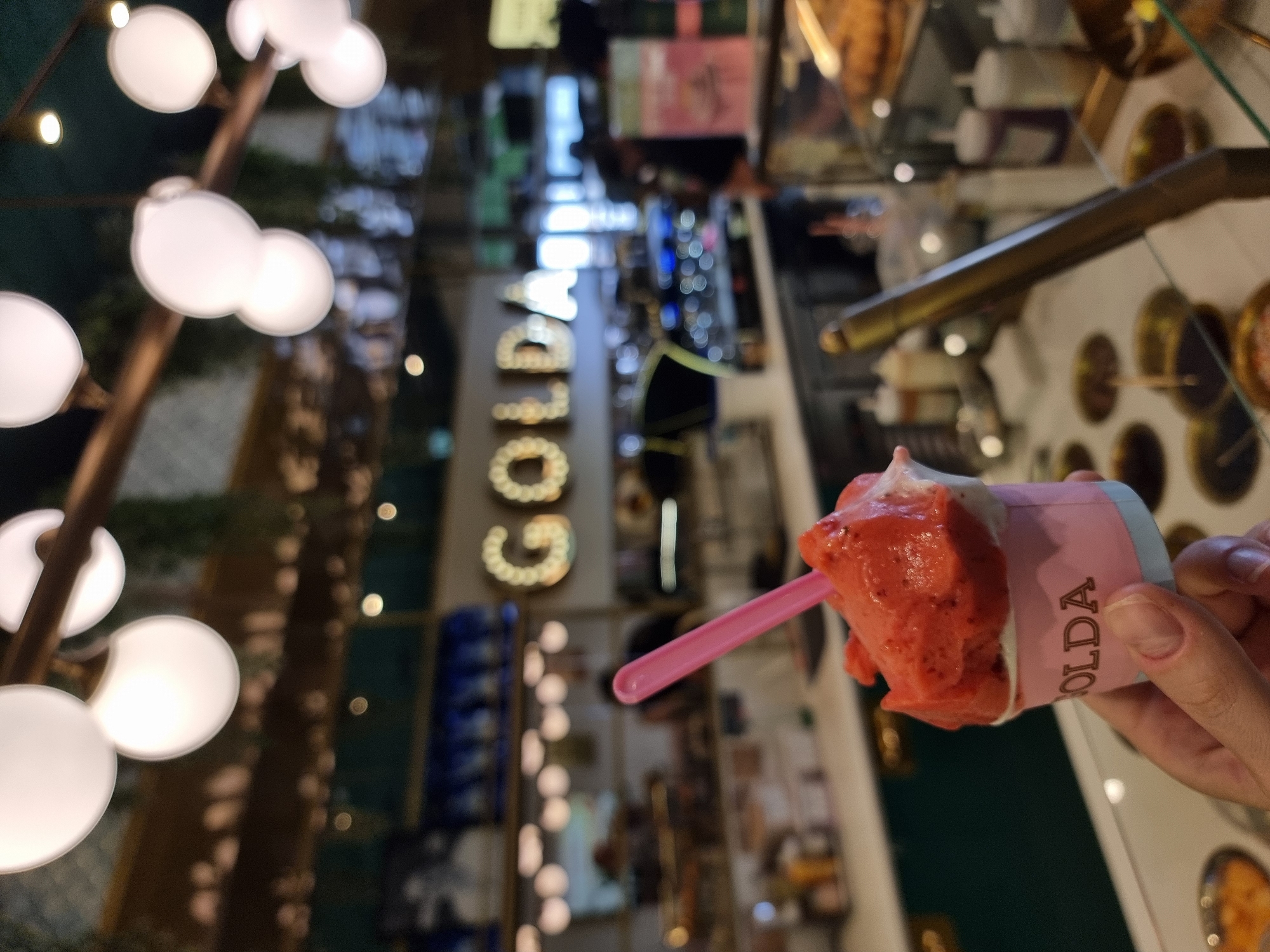 A close-up, canted image captures a cup of strawberry gelato held by a hand, with a pink plastic spoon inserted. The cup has the word "GOLDA" printed on it. The backdrop is a blurred interior of a gelateria, with visible display cases containing various flavors. Further back, the word "GOLDA" is spelled out in large, illuminated letters, matching the brand name. Soft, globular lights hang on the left. Based on the brand, this scene is likely taking place at a Golda ice cream shop in Bnei Shimon Regional Council, Israel.