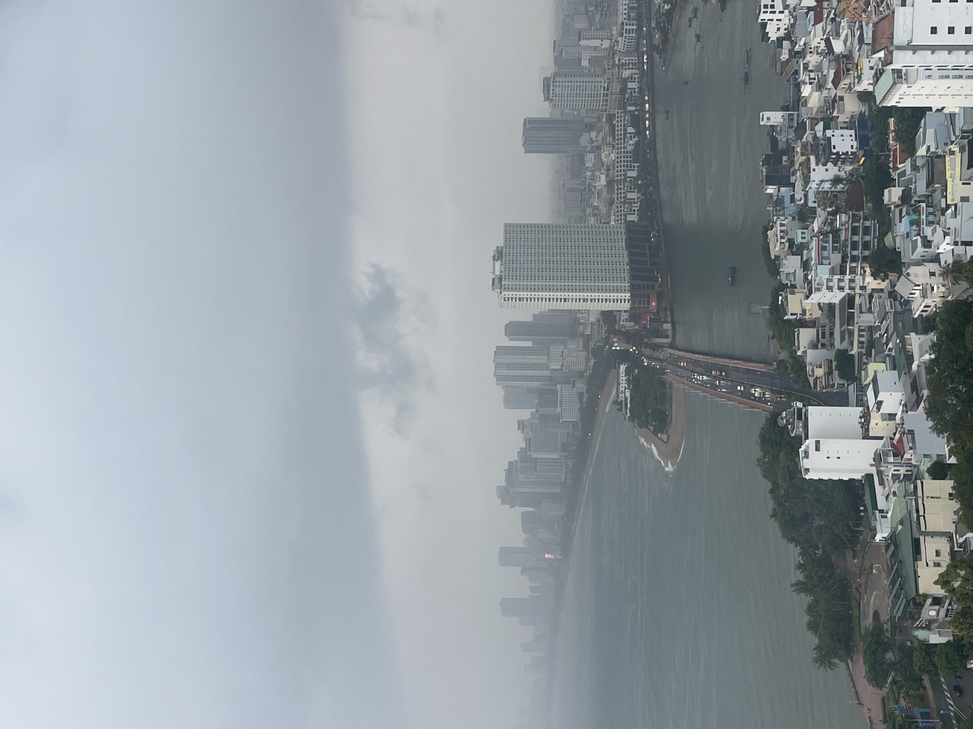 The image depicts an aerial view of a cityscape in Bac Nha Trang Ward, Vietnam, under heavy rain or fog, which significantly obscures details. A river flows vertically through the scene, separating a dense urban area on the right from a line of high-rise buildings on the left. A bridge with visible traffic connects both sides of the river. The buildings on the right are mostly low-rise residential and commercial structures, interspersed with trees. The sea is visible to the lower part of the frame.