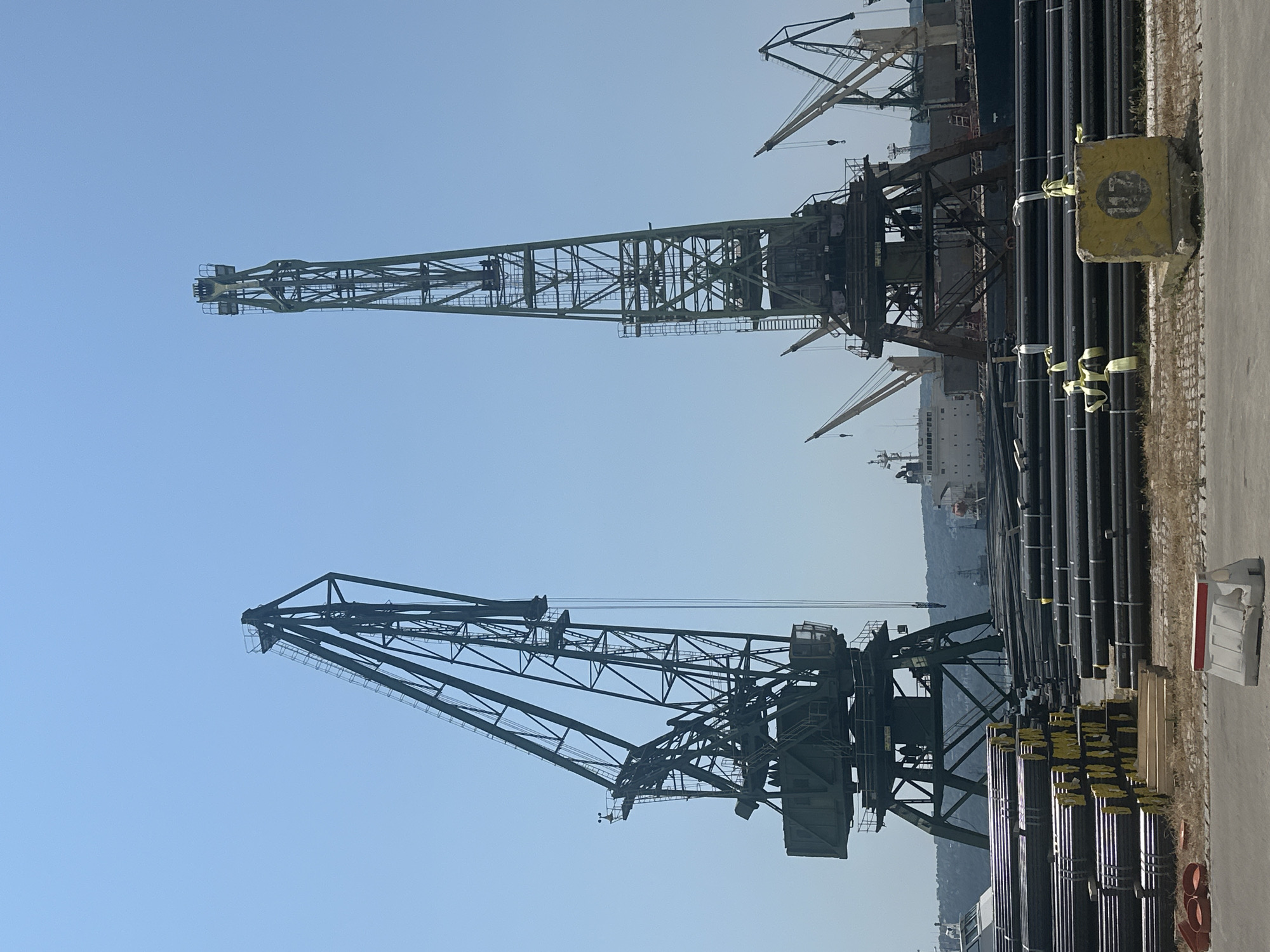 In the city of Unknown, Unknown, this image captures a view of a port on a clear day. Two large, green gantry cranes dominate the foreground, silhouetted against a bright blue sky. The crane on the left is positioned at an angle, while the one on the right stands taller and straighter. In the midground, a large cargo ship is docked, partially obscured by the cranes. Piles of long, black pipes are stacked in neat rows in the foreground, some bound with yellow strapping. Behind the pipes, other smaller cranes and industrial structures are visible, along with a hilly landscape in the distance. The ground is paved, with some dry grass and debris scattered around the pipe stacks. A yellow concrete block with the number "117" inscribed on it is visible in the lower right corner, and a white barrier with red markings is present in the lower left. The overall scene depicts a working industrial port environment.