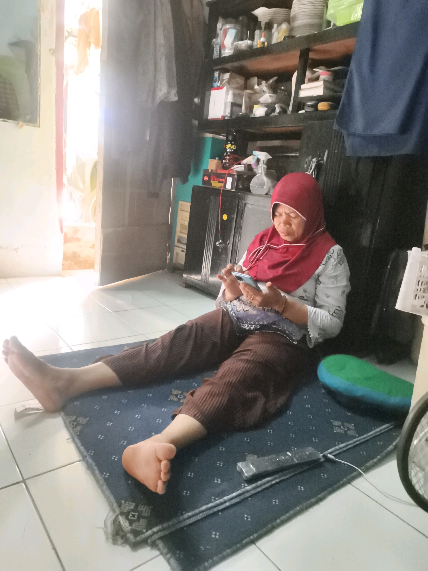 An adult woman, wearing a red hijab and a white and grey patterned shirt, sits cross-legged on a dark blue rug on a tiled floor. She is looking down at a blue mobile phone in her hands. She is wearing brown, ribbed trousers. Her legs are extended in front of her, and her feet are bare. To her right, a green and blue cushion is visible. In the background, shelves are stacked with various objects, including what appear to be dishes, containers, and bottles. A doorway with bright light emanating from it is visible to the left of the woman. A portion of a fan is visible on the far right of the image. The scene appears to be an indoor domestic setting, possibly a living room or a common area within a home in Cimahi, Indonesia.