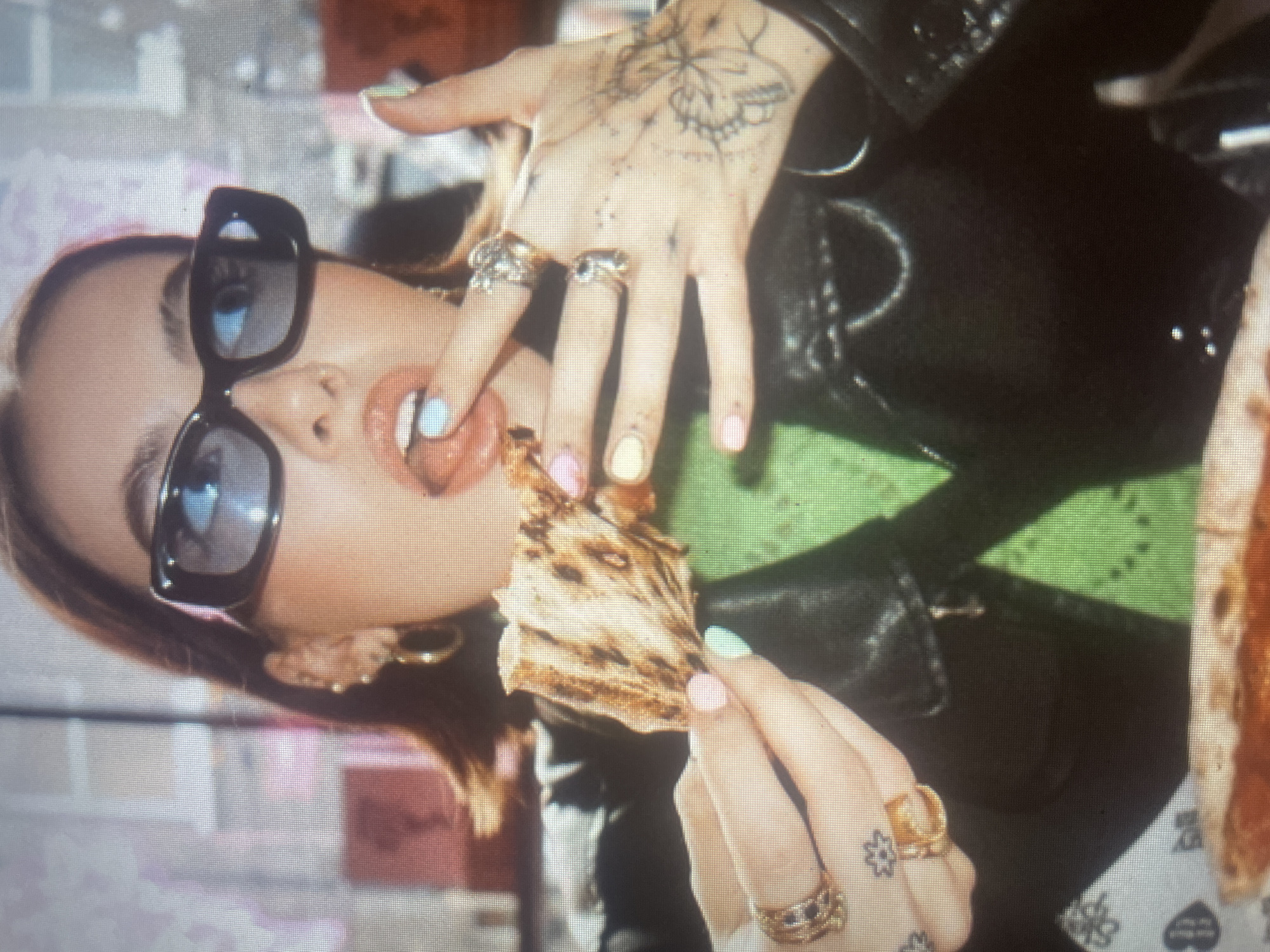 A woman with sunglasses is eating pizza in an indoor setting. She is wearing a black leather jacket and a green top. She has multiple rings on her fingers and tattoos on her hands. She is holding a slice of pizza and appears to be about to take a bite. The background is blurred but suggests a restaurant or public place.